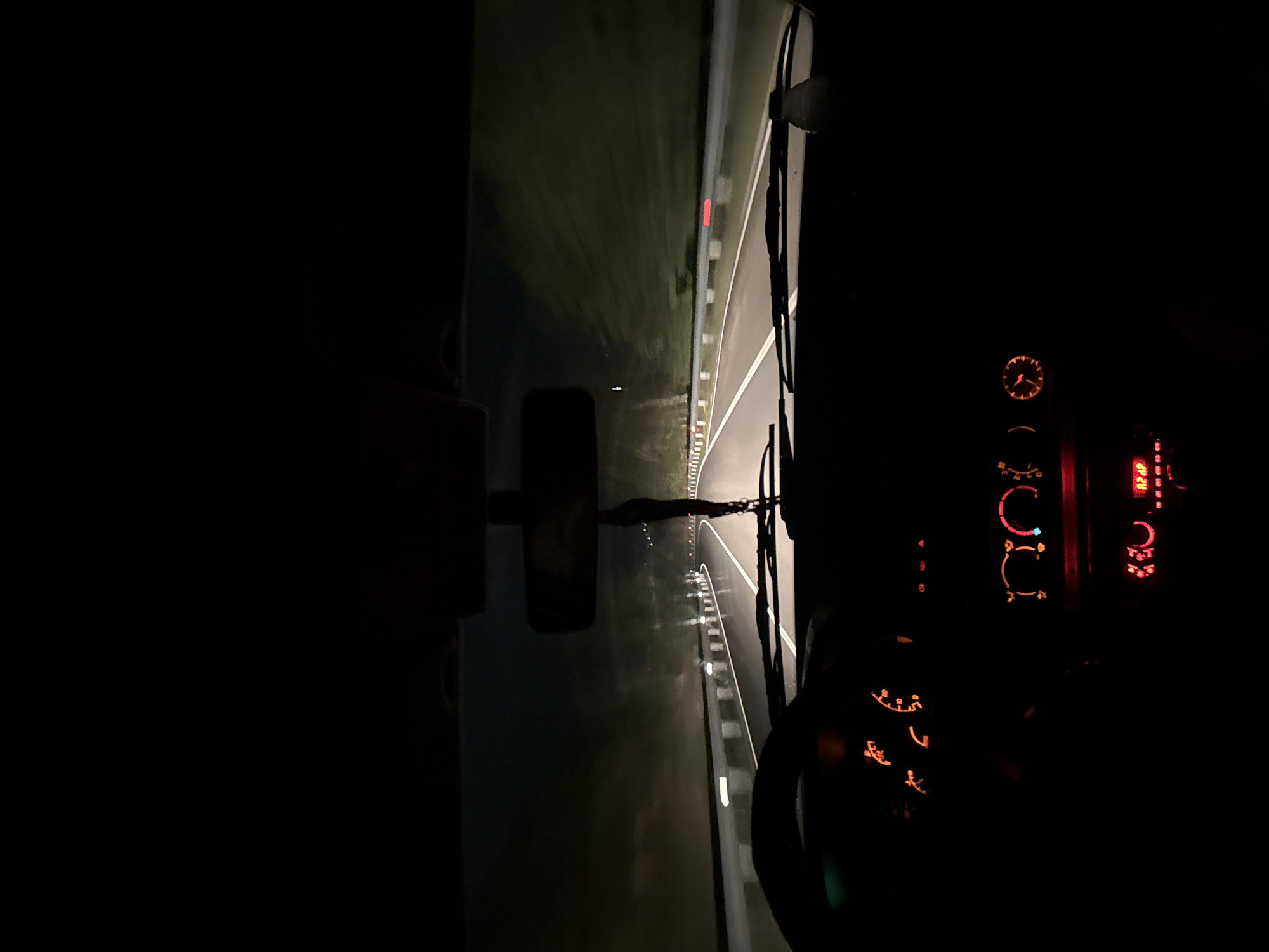The image captures a night-time scene from the interior of a vehicle, looking out through the windshield onto a road. The original photograph is oriented vertically but depicts a horizontal perspective.
Outside the vehicle, a dark asphalt road is illuminated by the vehicle's headlights, revealing clear white lane markings and a center line. To the right of the road, a metal guardrail stretches into the distance, punctuated by regularly spaced red and white reflectors. Beyond the guardrail, dark, grassy terrain is visible, with hints of faint, distant lights. A reflection of what appears to be a dark rectangular object, possibly a mobile phone, can be seen on the left side of the windshield.
Inside the cabin, the dashboard dominates the right side of the frame, aglow with a warm red and orange illumination from various gauges and controls. A small analog clock face with simple markings is visible at the top. Below it are several climate control dials, indicated by blue and red accents, along with an array of other illuminated buttons and a digital display showing partial alphanumeric text, possibly "R2HP". A windshield wiper arm is partially visible on the far right.
The scene depicts the experience of driving at night. The environment outside suggests a rural or semi-rural road, aligning with the location context of Sadovo, Bulgaria. The time of day is clearly night, with no indications of adverse weather. No individuals are visible, but the perspective implies the presence of a driver or passenger.