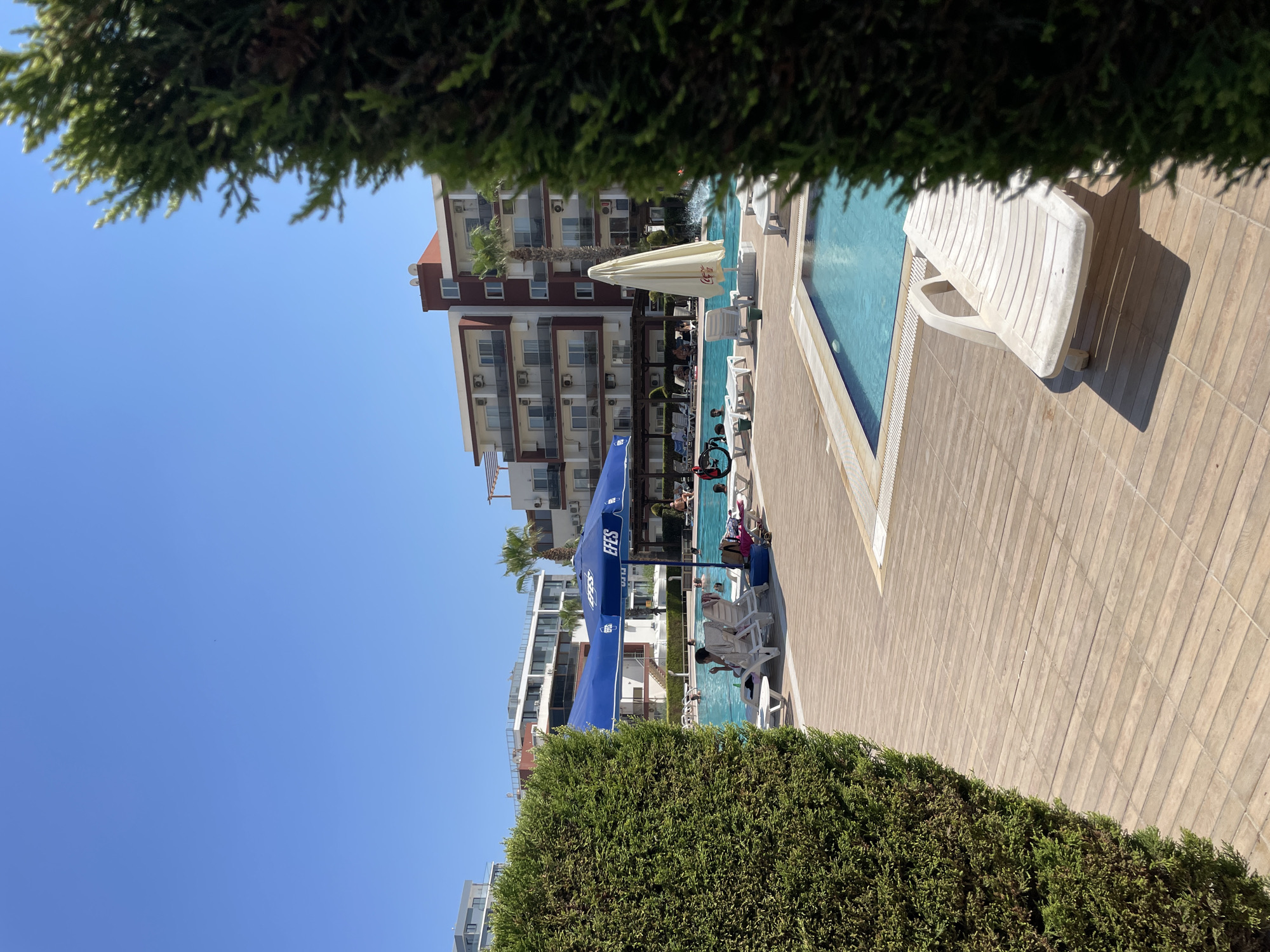 The image depicts a bright, sunny day at a resort in Trikomo, Northern Cyprus. A swimming pool with clear blue water is the central focus, surrounded by a wooden deck. Several lounge chairs are scattered on the deck, some occupied by people relaxing. Two large umbrellas, one blue with "EFES" branding and another white with "Coca-Cola" branding, provide shade. In the background, a multi-story hotel building with balconies and air conditioning units is visible, along with palm trees. Lush green bushes frame the foreground on both the left and right sides of the image. The sky is a clear, bright blue, indicating pleasant weather.