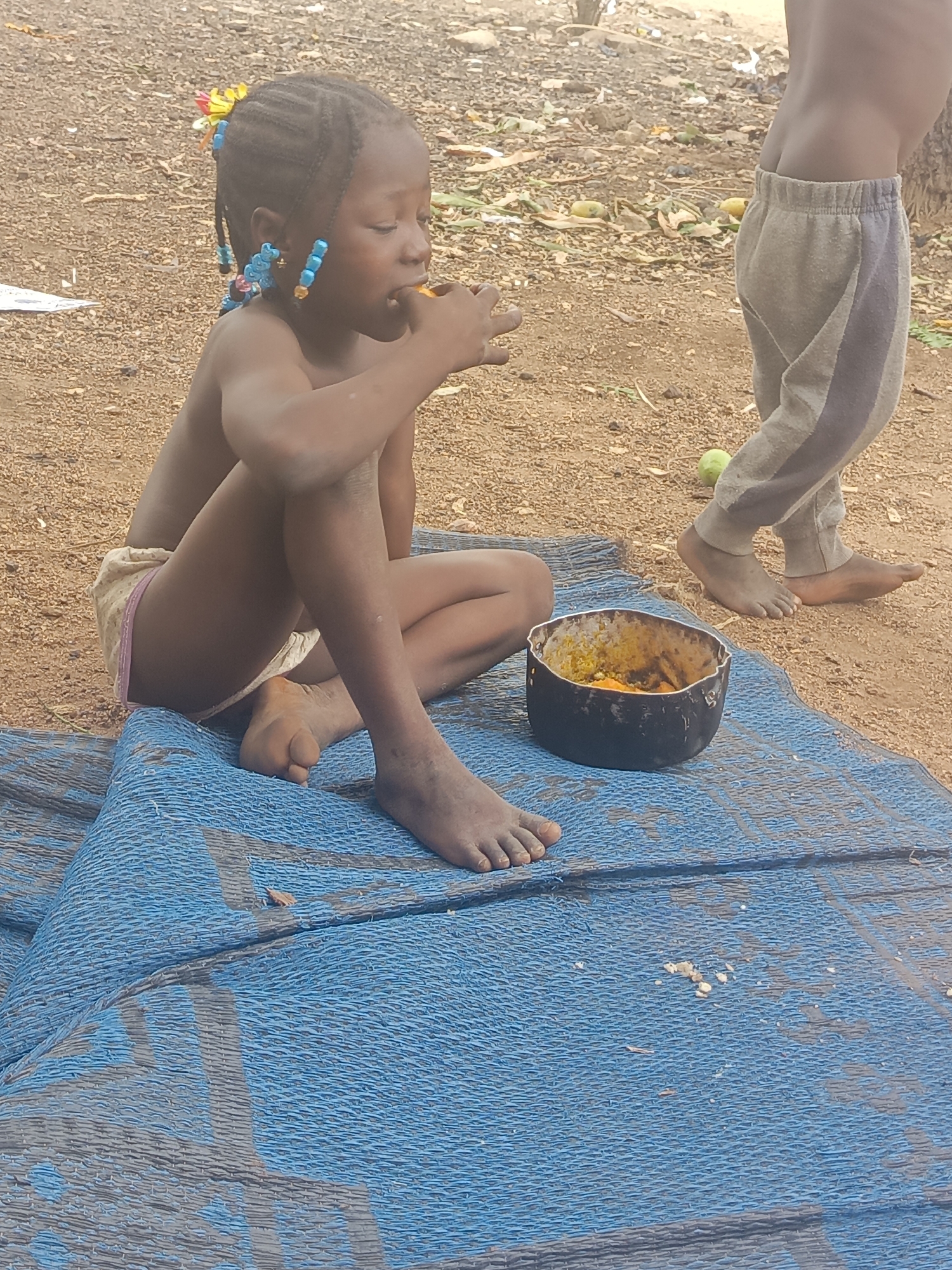 The image depicts a young, dark-skinned child eating from a dark-colored bowl. The child is sitting cross-legged on a blue and gray woven mat placed on reddish-brown ground with scattered debris. The child has braided hair adorned with colorful beads and flowers. Another child is partially visible in the upper right corner, wearing gray sweatpants and walking barefoot. The bowl contains a yellow-orange substance, presumably food. The environment appears to be an outdoor area. The photo was taken in the city of Akwanga, Nigeria.