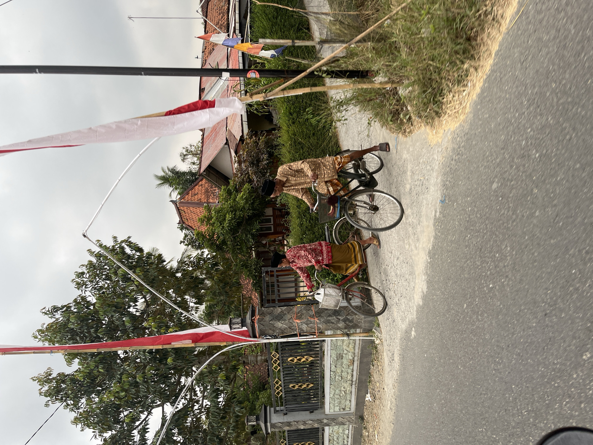The image captures a scene on a street in Karangtanjung, Indonesia, with two individuals on bicycles. A mature person in traditional Indonesian attire, including a red patterned shirt and a yellow sarong, is on a red bicycle with a basket in the front. They are wearing a black cap and sandals. Another person, also in traditional attire with a patterned brown shirt and a checkered sarong, is on a dark-colored bicycle. Both individuals appear to be moving forward on the paved road, which curves to the right. The background features a residential area with houses having red tiled roofs. Large trees with green foliage are visible, along with a fence and a stone pillar adorned with Indonesian flags. The sky is overcast.