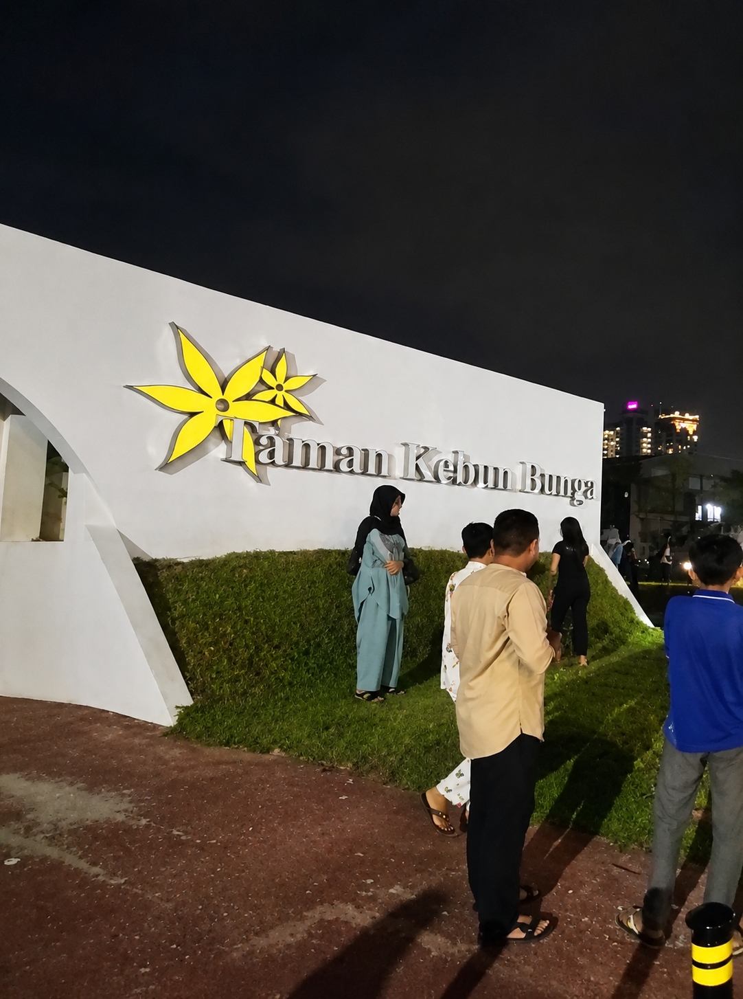 The image shows a nighttime scene at Taman Kebun Bunga in the city of Medan, Indonesia. A group of people are standing in front of a large white wall with the park's name, "Taman Kebun Bunga," in silver lettering and a yellow flower logo. The wall is illuminated, and a dark night sky is visible above. The ground in the foreground is a reddish-brown, paved surface, and a section of neatly trimmed green grass is to the right of the wall. Several individuals are present, including a woman in a light blue headscarf and dress, a person in a tan shirt and black pants, and two boys, one in a blue shirt and grey pants, and another partially visible. The overall atmosphere appears to be casual, possibly a public gathering or an evening visit to the park.