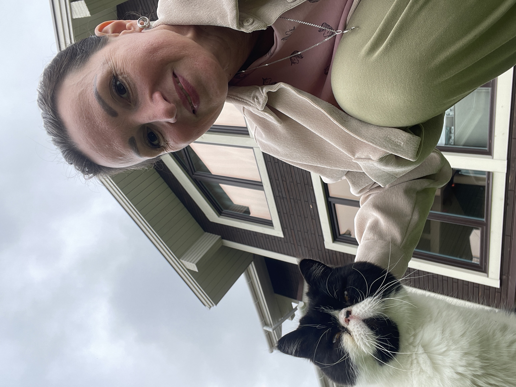 A medium shot captures a woman and a black and white cat in front of a two-story house with dark brown siding and white trim. The woman, appearing to be in her 40s, is crouched down and smiling at the camera, her face illuminated by natural light. She is wearing a light pink t-shirt with a subtle pattern, a beige jacket, and olive green pants. A silver necklace with a pendant rests on her shirt. The cat, a domestic shorthair with distinct black and white markings, is positioned to the left of the woman, looking directly at the viewer. Its fur appears fluffy, and its whiskers are prominent. The background consists of the house's facade, featuring multiple windows with a reddish tint, and a cloudy sky. The perspective is low-angle, emphasizing the subjects from a ground-level viewpoint.