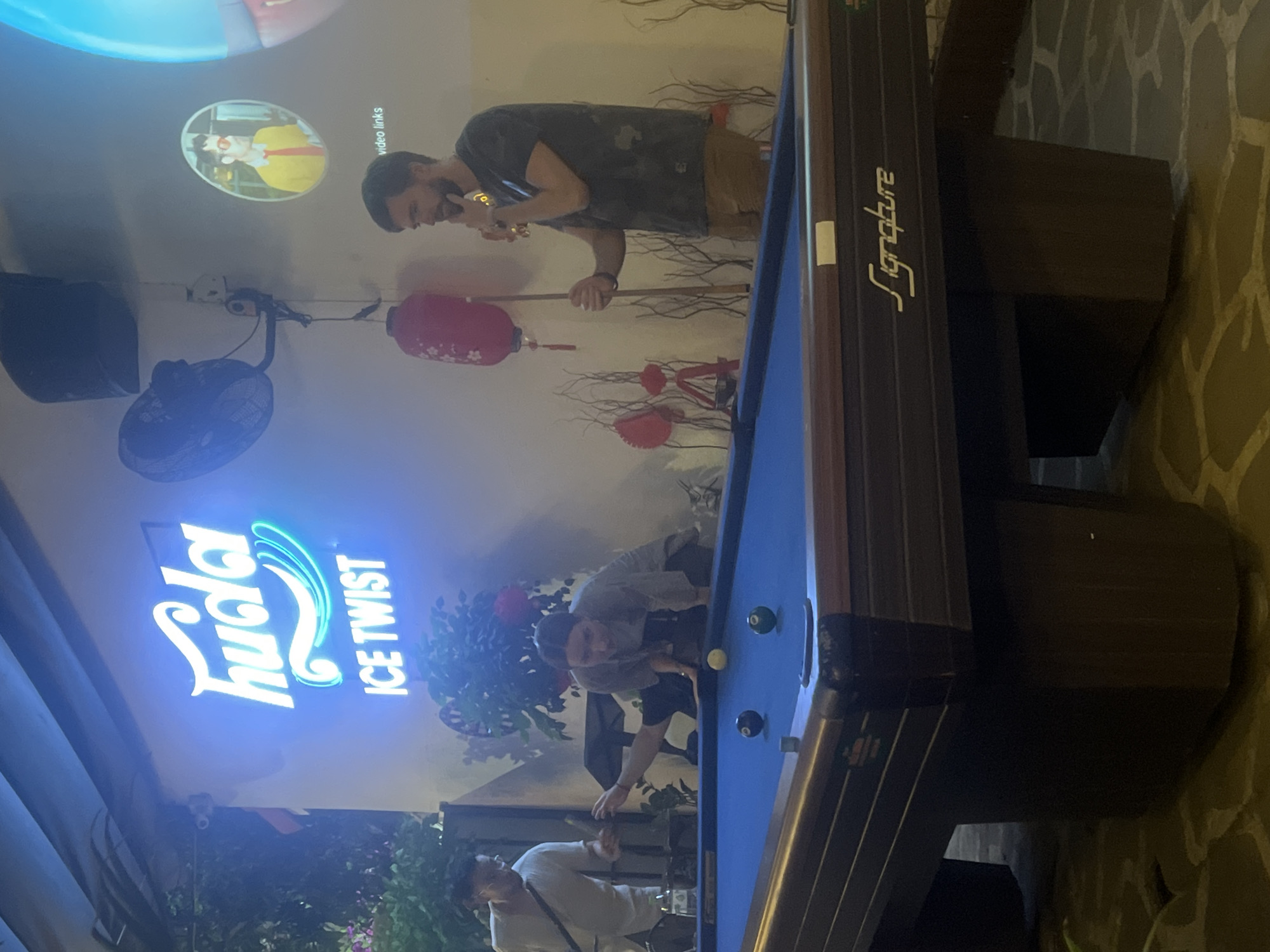 This photo, taken in Đà Nẵng, Vietnam, depicts a nighttime scene at what appears to be a bar or entertainment venue with Vietnamese cultural influences in decor. The location features a "Huda Ice Twist" neon sign, indicating the sale of Huda beer. A pool table occupies the right side of the frame, with several billiard balls visible. Two men are playing or preparing to play pool. One man is leaning over the table, seemingly taking a shot. Another man in a dark t-shirt is standing, holding a cue stick, and talking on the phone. A red lantern and tree branch decorations adorn the wall. A video is playing on a screen above the pool players. A ceiling fan is attached to the wall and turned on.