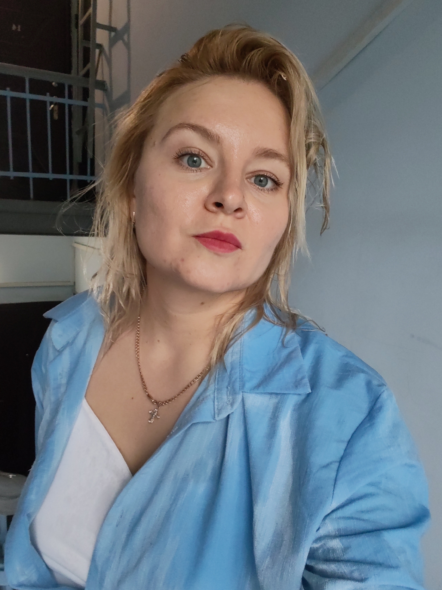 The image is a close-up, waist-up selfie of a fair-skinned woman with blonde hair and blue eyes. She is wearing a light blue collared shirt over a white top and a gold chain necklace with a cross pendant. Her lips are painted with a bright pink lipstick. The background consists of a light blue wall on the right and a dark, industrial-looking structure with a railing on the left. There is no discernible information in the image that would suggest a specific location within the city of Izhevsk, Russia. The woman appears to be indoors.