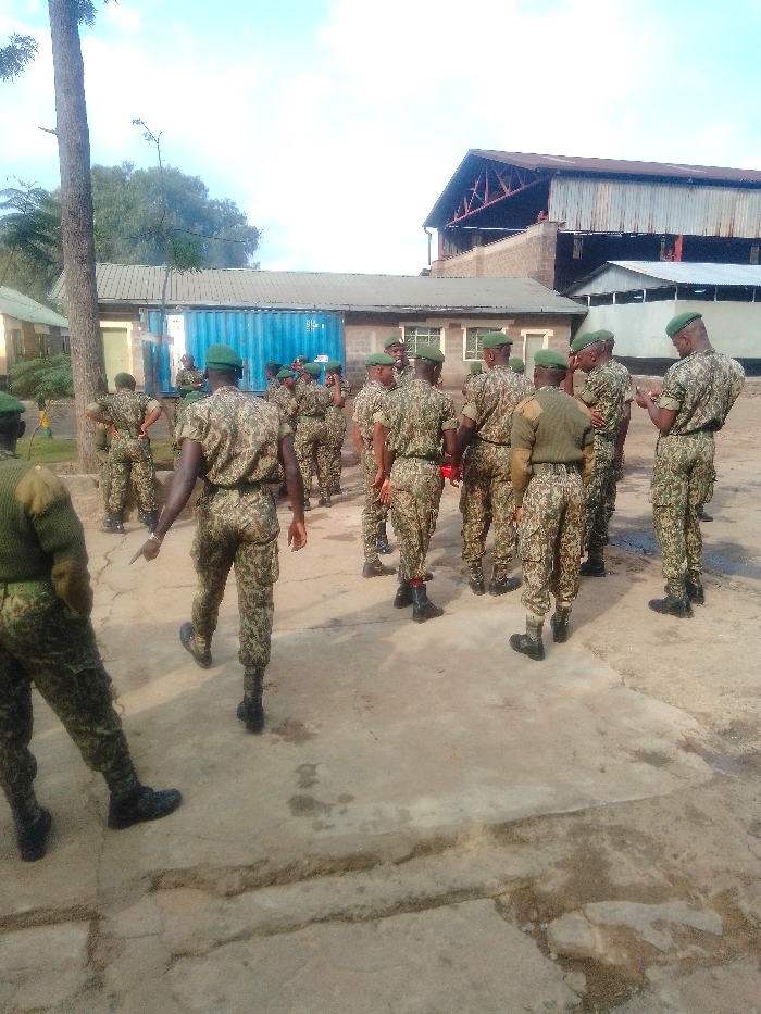 The image depicts an outdoor scene at an unspecified military or training facility in Nairobi, Kenya. Approximately 20-25 adult males, uniformly dressed in green and brown camouflage fatigues and green berets, are present. They are primarily standing on a cracked, light-grey concrete surface that exhibits various stains and wear.
One individual in the foreground, facing away from the camera, is walking towards the left side of the frame and pointing with his right index finger. The other individuals are scattered in loose clusters across the area; some appear to be conversing, while one on the far right is observed looking down at an object held in his hands.
In the mid-ground, a prominent, long blue shipping container is situated, likely repurposed for storage, with several single-story buildings visible behind it. These buildings feature light-colored walls and dark, possibly corrugated, roofs. To the far right, a large industrial-style building is visible, characterized by a reddish-brown corrugated metal roof supported by exposed metal framework and dark walls below. A tall, slender tree trunk rises on the left side of the frame, with additional green foliage visible in the distant background. The sky overhead is clear and light blue with minimal cloud cover, indicating daylight conditions.