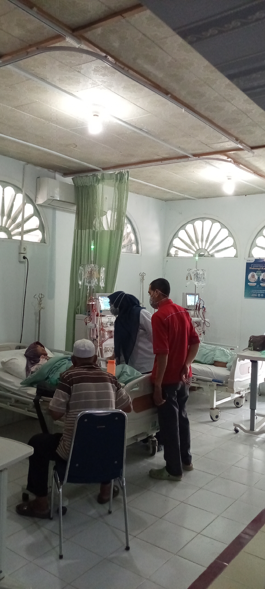This is a medium shot, indoor, vertical photograph taken in what appears to be a medical facility, possibly a dialysis center or a ward in a hospital. The room has white tiled floors and walls, and a textured, drop-ceiling with exposed electrical conduits and fluorescent lighting. Large, arched windows with decorative pane patterns are visible on the left and right sides of the room, letting in natural light. A green curtain hangs centrally, dividing some of the space and obscuring a portion of the room.
In the foreground, a man wearing a white and brown striped shirt, dark pants, and a white cap is seated on a dark blue chair, facing away from the camera. To his right, two individuals in medical attire are attending to a patient in a hospital bed. One individual, wearing a dark hijab and a white scrub top, is looking at a medical monitor displaying readings. The other individual, wearing a red short-sleeved shirt and dark pants, stands beside them, also appearing to be observing the patient or the equipment.
In the background, two hospital beds are visible. One bed on the left has a patient lying in it, covered with a light green blanket. The other bed on the right is mostly obscured. Several medical equipment stands with IV bags and tubing are positioned around the beds, along with dialysis machines. A poster with Indonesian text and images is mounted on the far right wall. The overall impression is one of a clinical environment where medical care is being administered. The location is identified as Lhokseumawe, Indonesia.