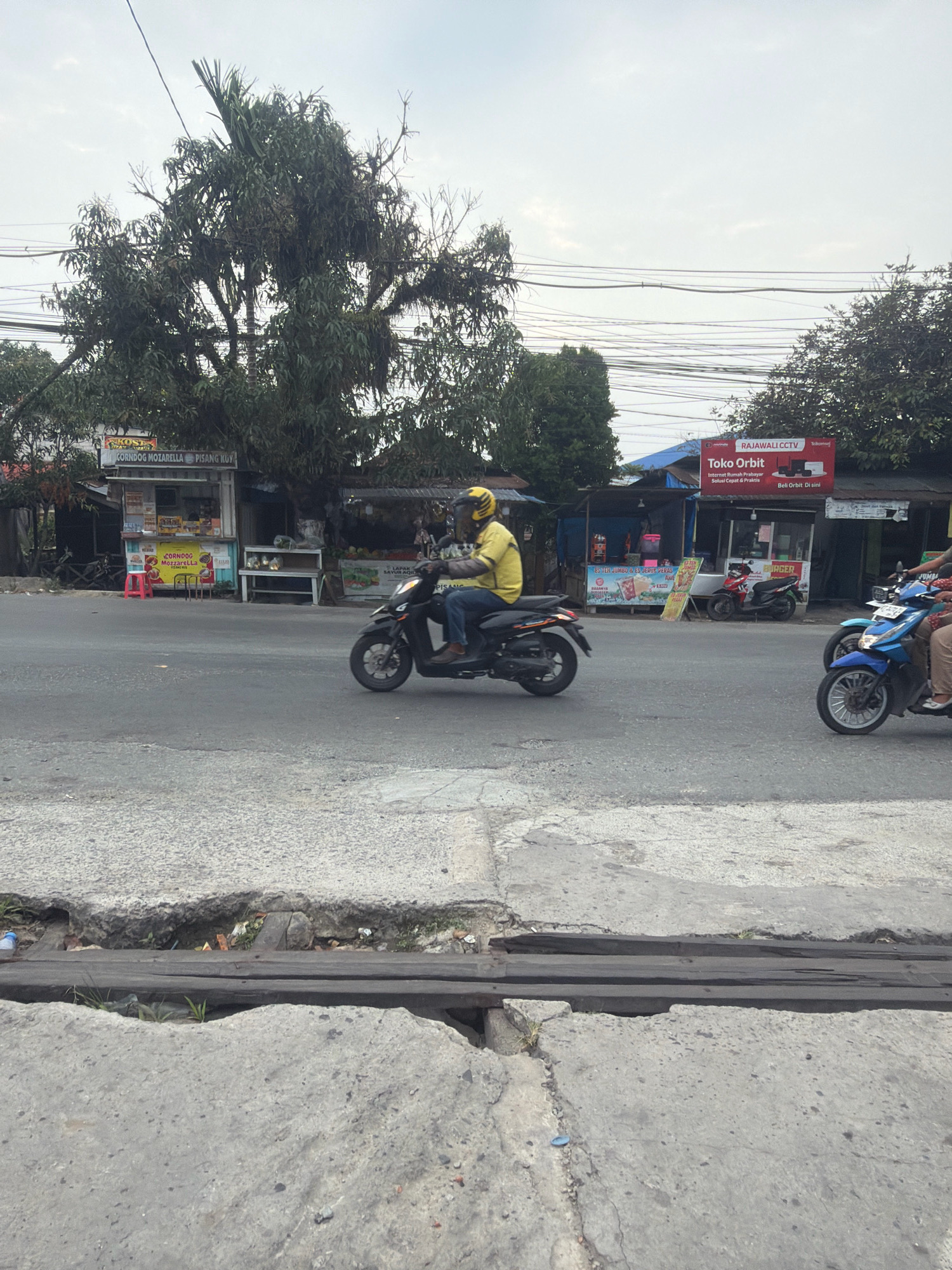A man in a yellow shirt and helmet rides a black scooter on an asphalt road in Balikpapan, Indonesia. He is in the left lane, moving forward. Several small roadside shops are visible on the right side of the road, featuring signs for food and services like "CORNDOG MOZZARELLA," "ES TEH JUMBO," and "Toko Orbit RAJAWALI CCTV." There are power lines overhead. Two other scooters, one blue and one possibly red, are parked or stopped near the shops. The road has visible cracks and a drainage grate. Trees and vegetation are present in the background. The sky is overcast.