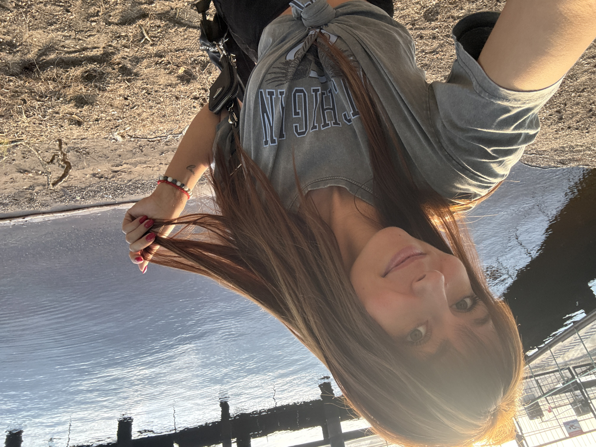 A young woman with long brown hair and bangs is taking a selfie by a body of water. She is wearing a gray t-shirt with a graphic print and black pants, and she is holding a strand of her hair. The background shows a pier or dock on the left and calm water reflecting the sky and structure. The ground in the foreground is sandy and dry with some sparse vegetation. The lighting suggests it is late afternoon or early morning with the sun casting a warm glow. The location appears to be outdoors near a waterfront area, possibly a public park or recreational spot.