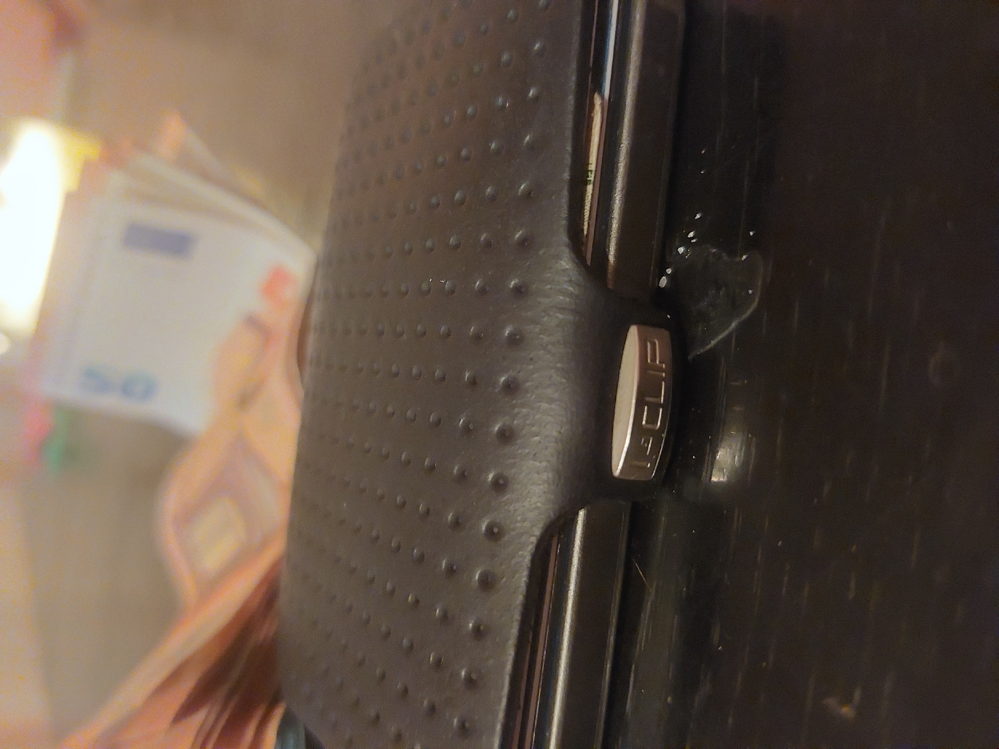 A close-up shows a black leather wallet with a textured surface and a metal clasp labeled "CLIP". Partially visible to the left of the wallet are blurred Euro banknotes, with a "50" visible on one of them. The setting or location is not discernible from the image.