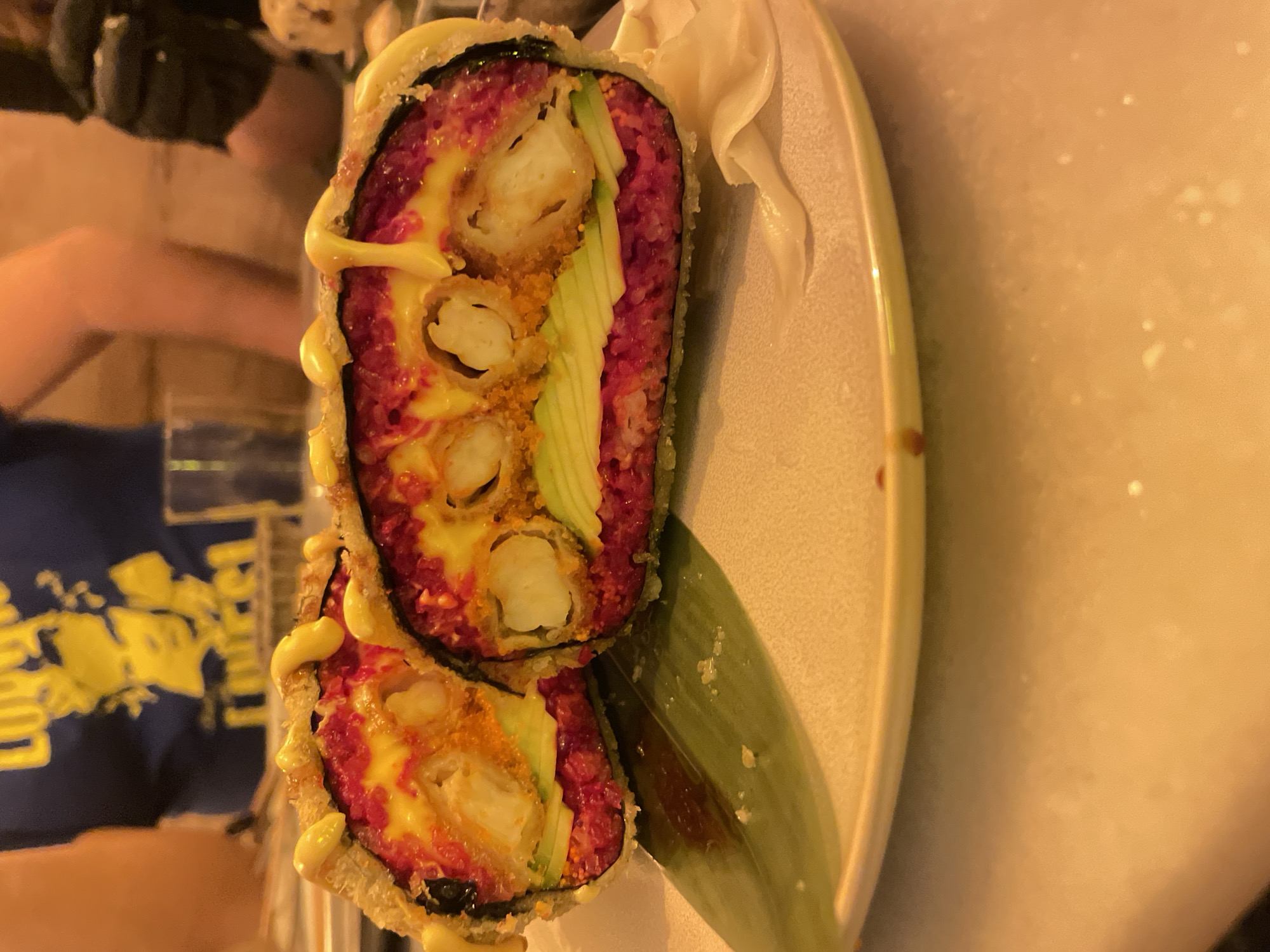A plate of two pieces of sushi lies on a white plate. The sushi is covered in a red rice and fried batter. The pieces are filled with a white filling, avocado slices, and a yellow sauce. One piece of sushi is stacked on top of the other. A green leaf is placed under the sushi. In the background, two people can be seen, one wearing a dark blue shirt with yellow writing. The location is believed to be Barcelona, Spain.