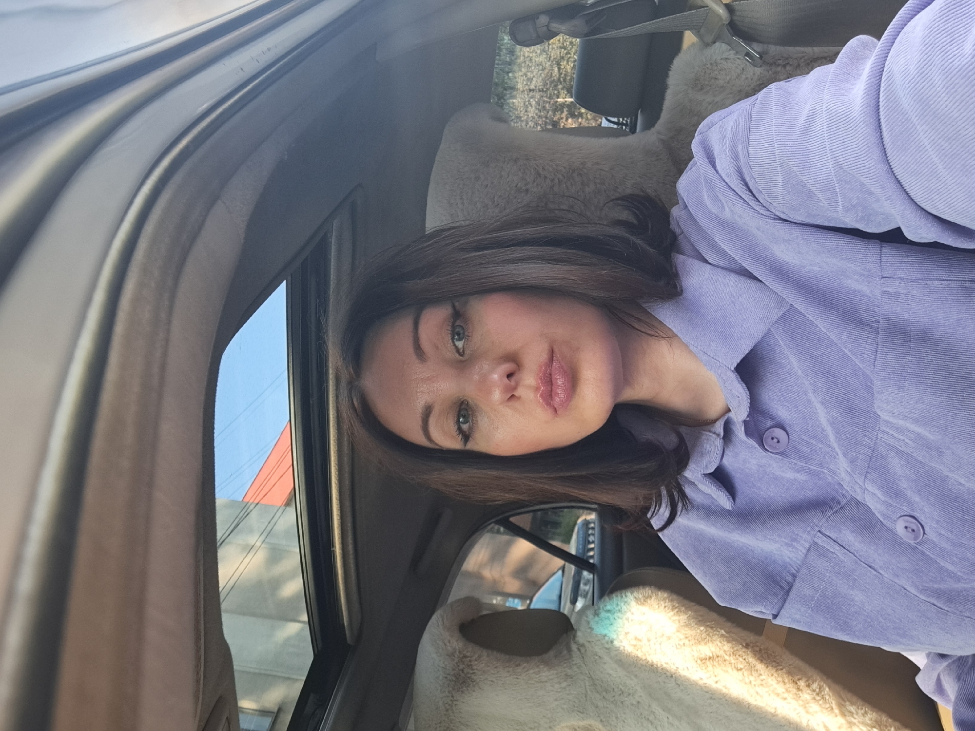 The image shows a woman with brown hair and makeup, wearing a light purple corduroy shirt, sitting in the passenger seat of a car. The car has a sunroof, which is open, revealing a building and the sky. The woman is looking directly at the camera with her lips pursed. The interior of the car is visible, including the fuzzy beige seat covers. The location is identified as Irkutsk, Russia, but there are no specific visual cues in the image to confirm this.