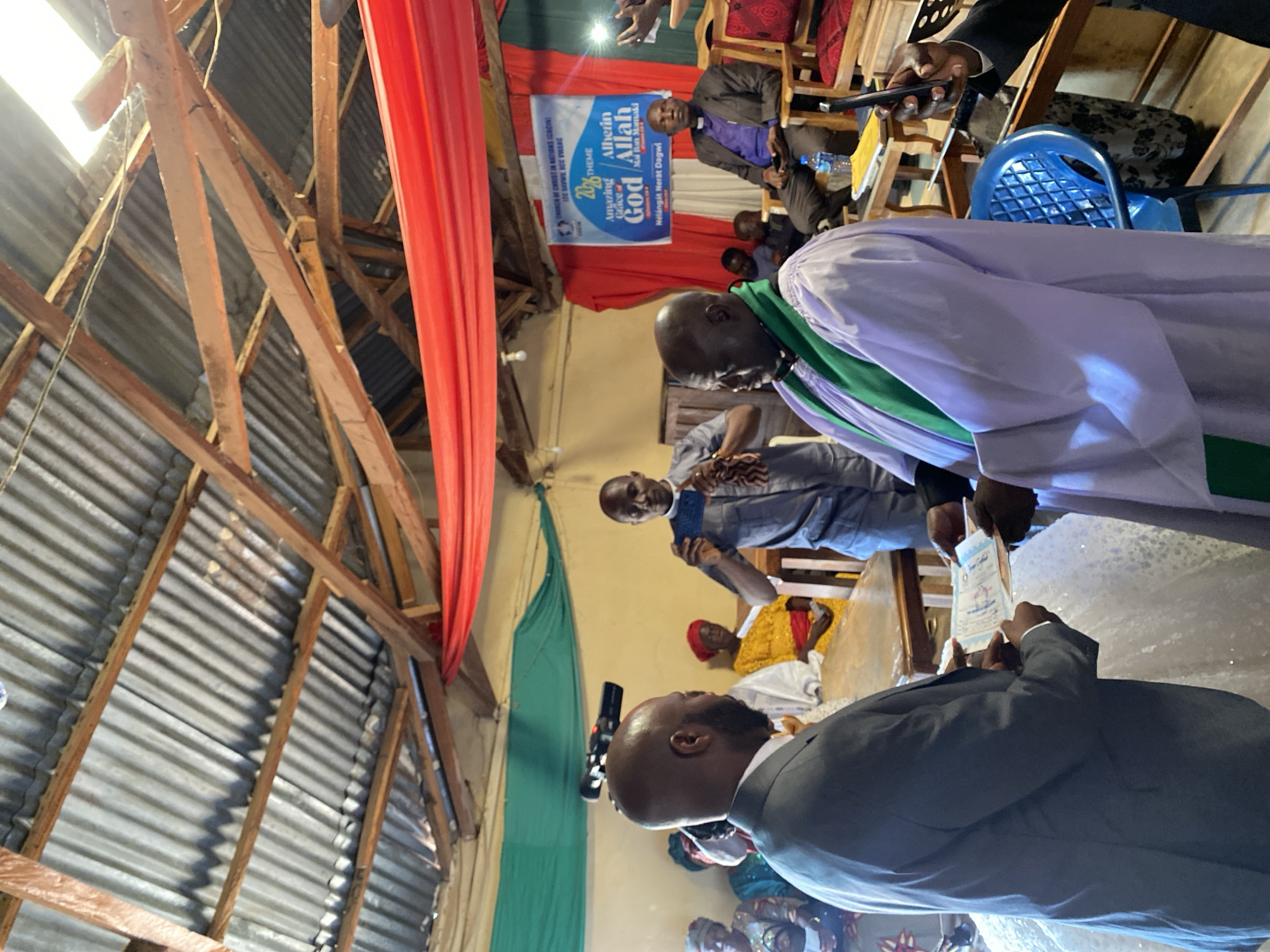 A view inside the Church of Christ in Nations (COCIN) in Vom, Nigeria shows a group of people gathered. Some are seated, including a man wearing a purple shirt and a black suit jacket and another wearing a white robe with green trim. A man in a gray suit stands with his back to the camera, holding a book or card. There is a banner behind the seated man that reads "Amazing Grace of God, Alherin Allah" and includes the church name and the text "2026 theme". Red and green curtains are hung around the perimeter of the room. The building has a roof made of corrugated metal, supported by wooden beams.