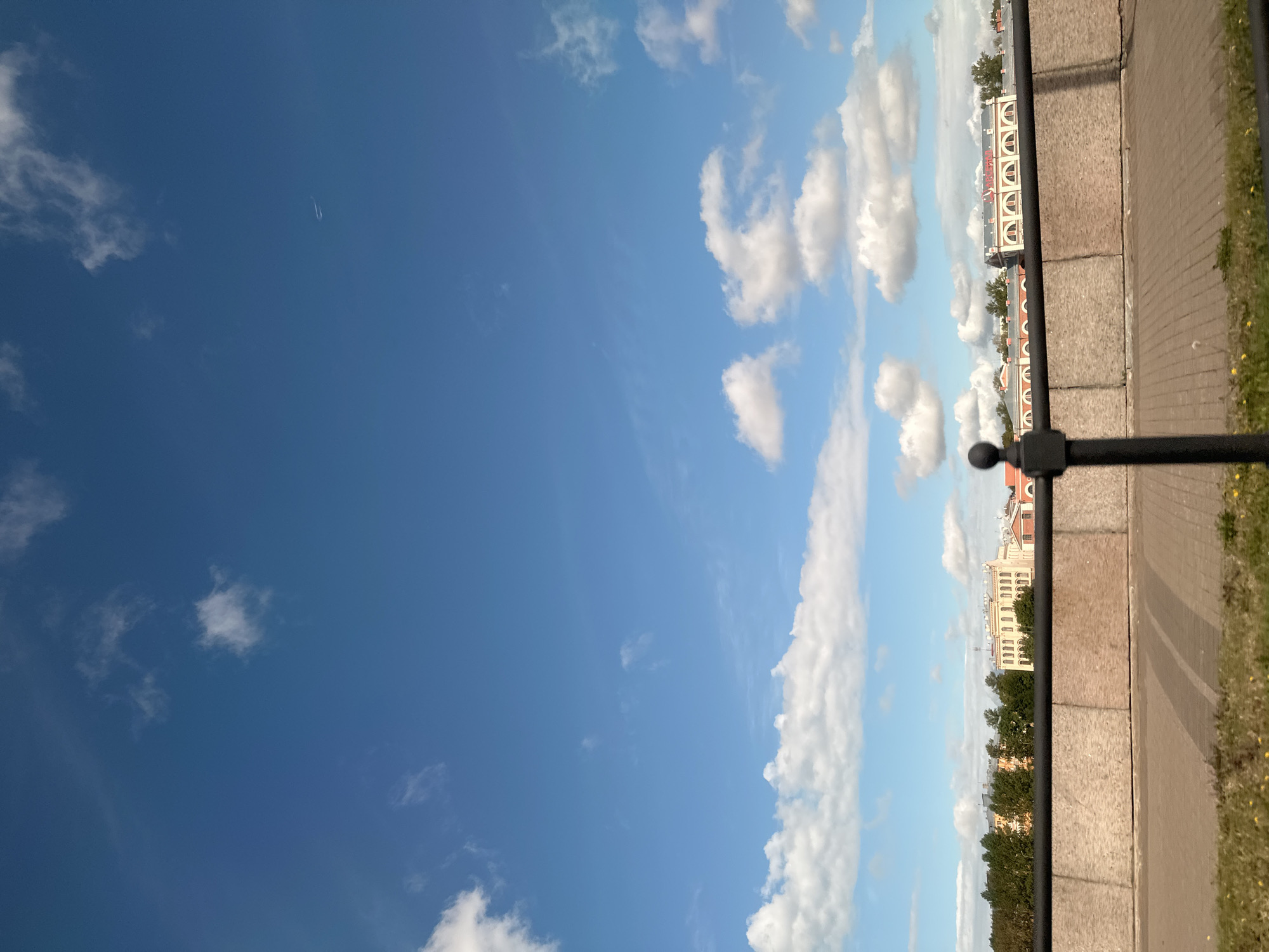This is a photograph taken from a quay in Saint Petersburg, Russia, likely during the daytime under a blue sky with scattered white clouds. In the foreground, a dark metal railing with a spherical finial runs horizontally across the frame, partially obscuring a stone embankment and a paved walkway. Beyond the embankment, a line of trees separates the quay from a row of light-colored, multi-story buildings with arched windows. The architecture suggests a historical or institutional setting, possibly along a canal or river. A faint contrail from an aircraft is visible in the upper right quadrant of the sky. The overall scene depicts a peaceful urban landscape with a clear sky.