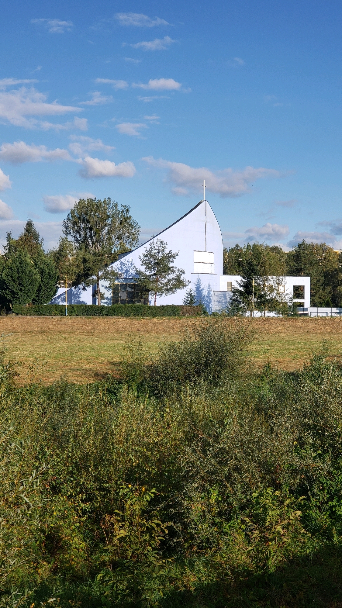 The image depicts the modern facade of the Church of the Holy Spirit in Prešov, Slovakia, situated amidst a lush green landscape under a bright blue sky dotted with white clouds. The church features a striking, curved roofline culminating in a cross at its apex, painted in a pale blue hue. A large, dark-paneled window is visible on the left side of the building, reflecting the surrounding trees. To the right, a series of white rectangular structures and windows break up the building's façade. The foreground is dominated by dense, verdant foliage and tall grasses, suggesting a natural, somewhat wild setting. The mid-ground consists of a dry, grassy field with scattered trees, leading up to the church complex. The overall impression is one of contemporary architecture harmoniously integrated into a natural environment. There are no people visible in the image.