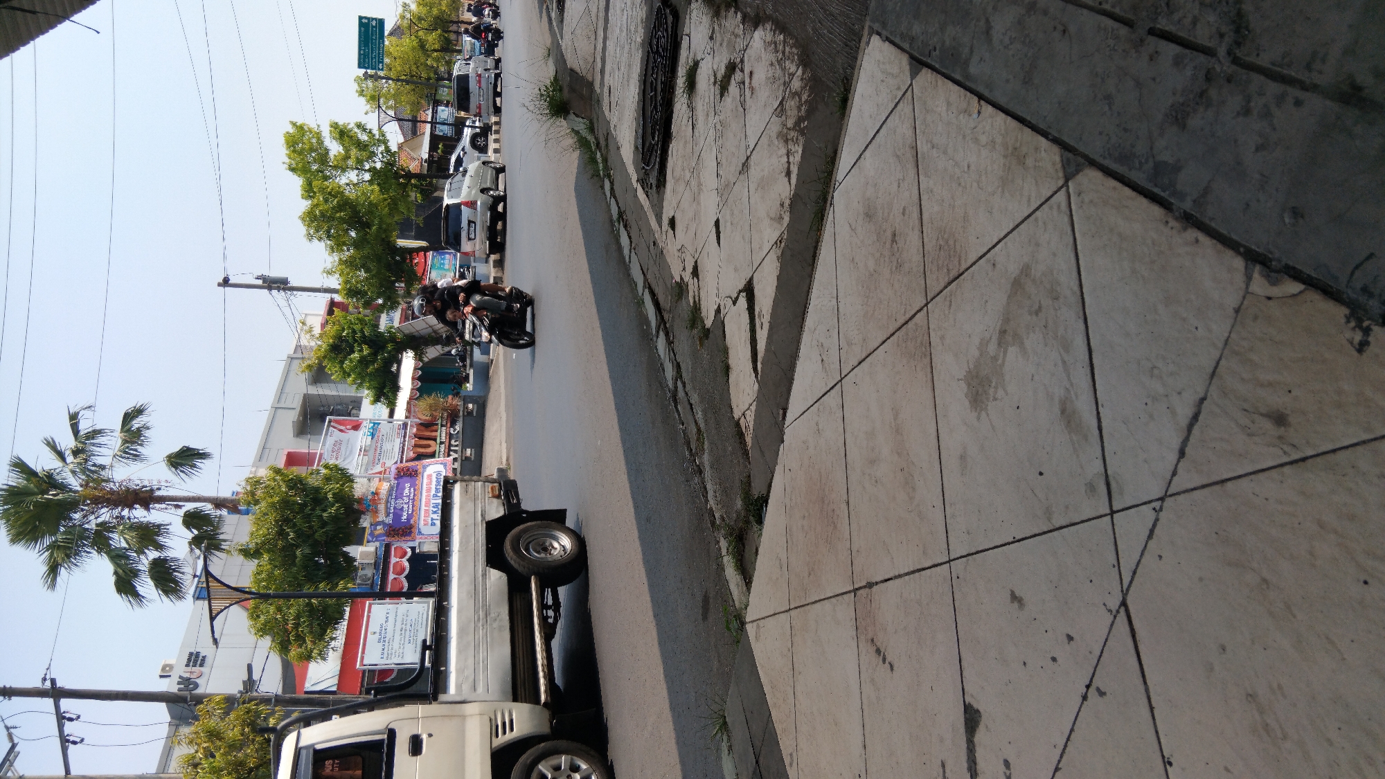 This image captures a street scene in Batang, Indonesia, likely on a sunny day given the bright lighting and shadows. In the foreground, a sidewalk paved with square concrete tiles is visible, showing some wear and tear with stains and cracks. A curb separates the sidewalk from the road.
The road is asphalt and shows signs of traffic. Several vehicles are present: a white pickup truck is parked or moving slowly on the left side of the road. Further down the road, a motorcycle with two riders is in motion, with the rider wearing a helmet. Behind the motorcycle, a silver car and a white van are also on the road, along with other vehicles in the distance.
The background features buildings, some of which appear to be commercial establishments. Banners and signs are visible on these buildings, suggesting a commercial or public area. Palm trees and other greenery are interspersed, indicating a tropical or subtropical climate. Overhead, electrical wires and poles are prominent. The sky is clear and blue. The overall impression is of a typical urban street in Indonesia, with active traffic and commercial activity.