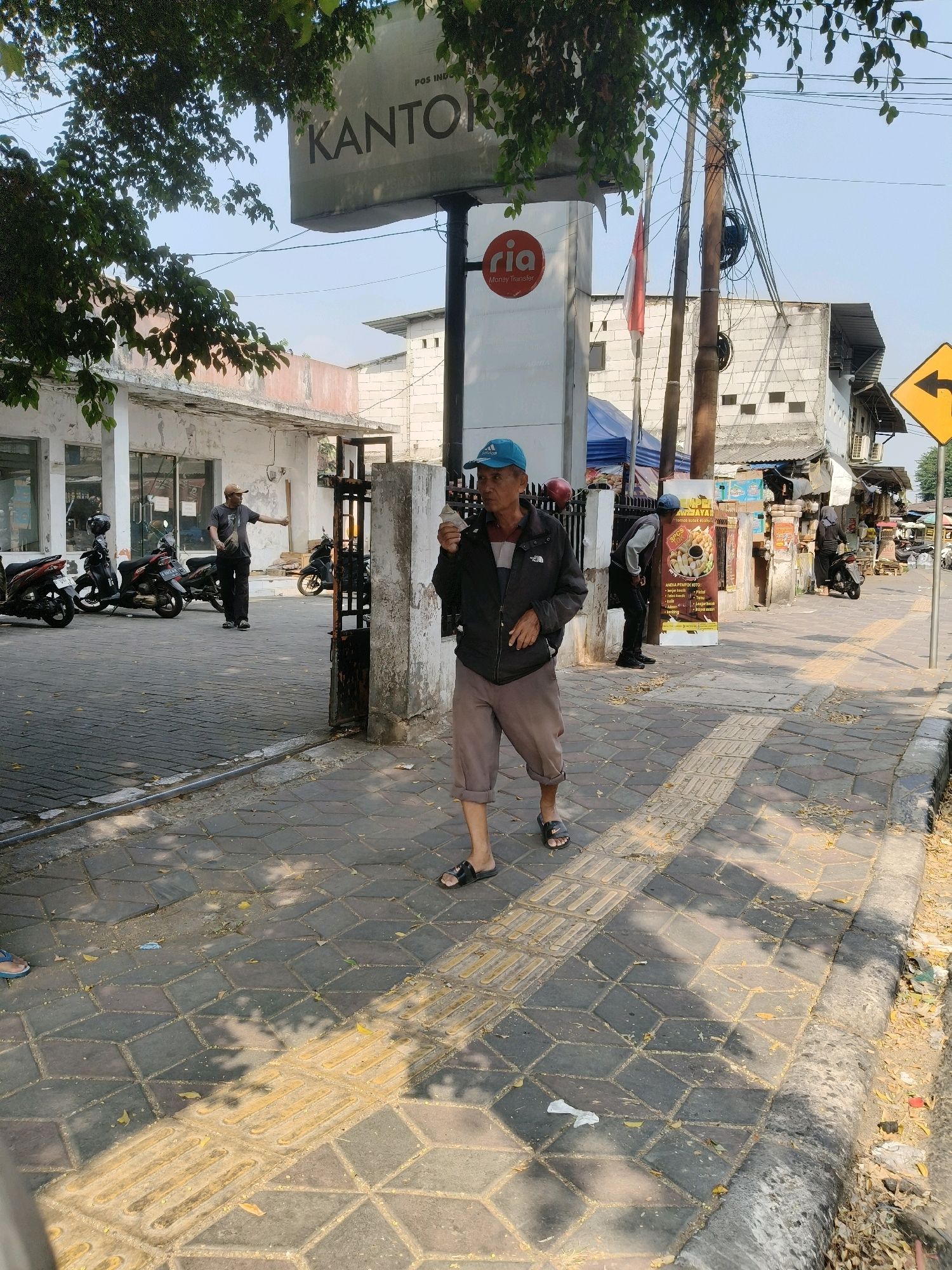 The image depicts a street scene in South Tangerang, Indonesia. In the foreground, a man wearing a blue baseball cap, a black jacket, and light brown shorts walks on a patterned brick sidewalk. He is holding a small white object in his right hand, possibly a piece of food. To his left, a tactile paving strip runs parallel to the curb. In the mid-ground, a man in a brown cap and dark t-shirt stands near a metal gate, gesturing with his right hand. Several motorcycles are parked in a lot behind him. A sign above them reads "KANTOR POS INDONESIA". To the right of the gate, a large banner with text and images of food items is visible. Further down the street, more people and vehicles are present, along with buildings and some market stalls. The environment appears to be a commercial or public area, with buildings lining the street and evidence of pedestrian and vehicle activity. Overhead, trees with green leaves cast dappled shadows on the ground. The sky is clear and blue.