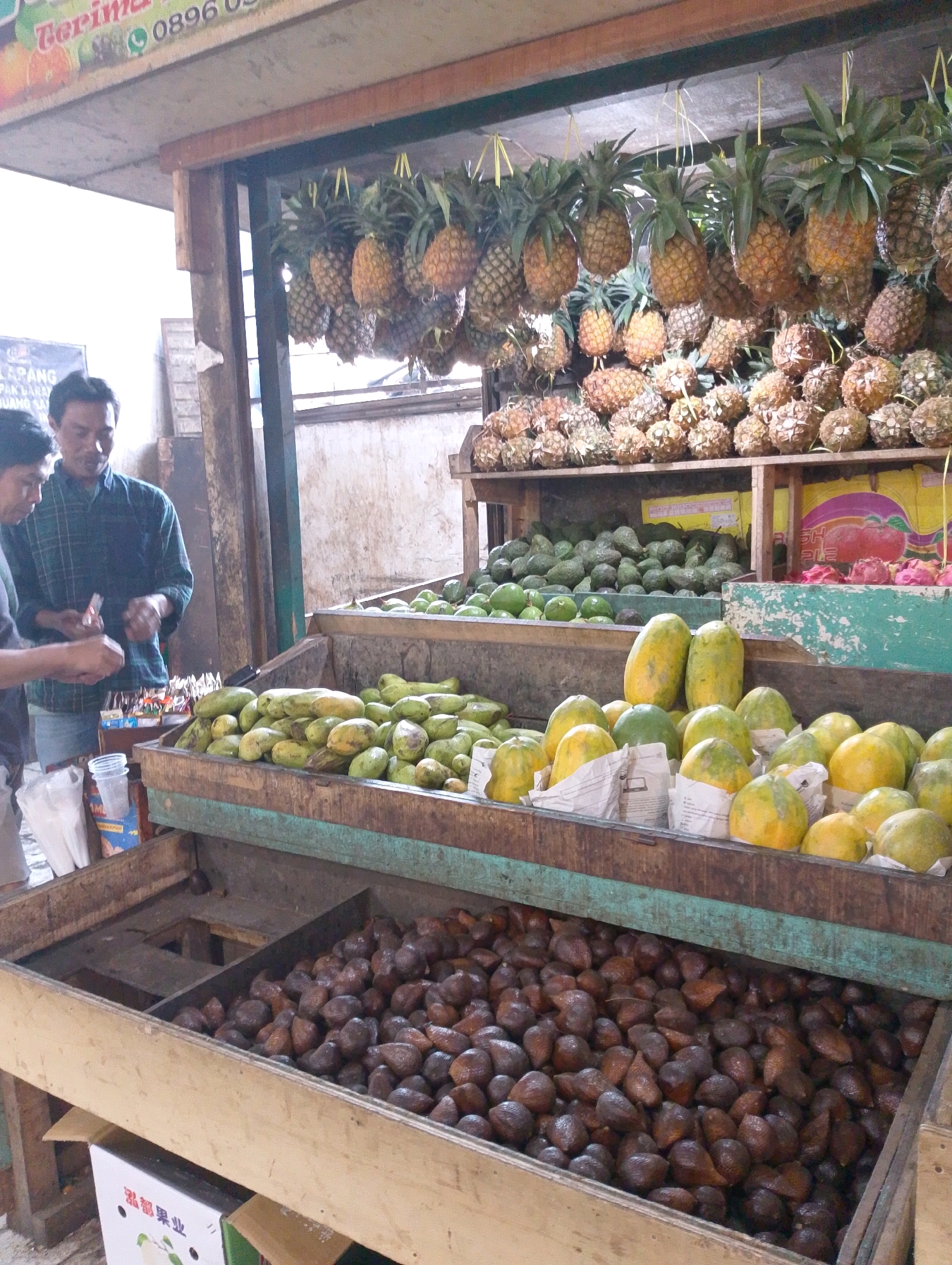 The image depicts an outdoor fruit stall in Lembang, Indonesia, a popular tourist destination known for its agricultural products. The stall features a wide variety of fresh fruits, with pineapples hanging from the ceiling and other fruits like papayas, avocados, and salak (snake fruit) displayed in wooden crates. Two men are present, one appears to be a vendor interacting with a customer, exchanging what looks like money and a small package. The background shows a rustic wooden structure, typical of traditional Indonesian markets. The overall scene captures the vibrant atmosphere of a local fruit market.