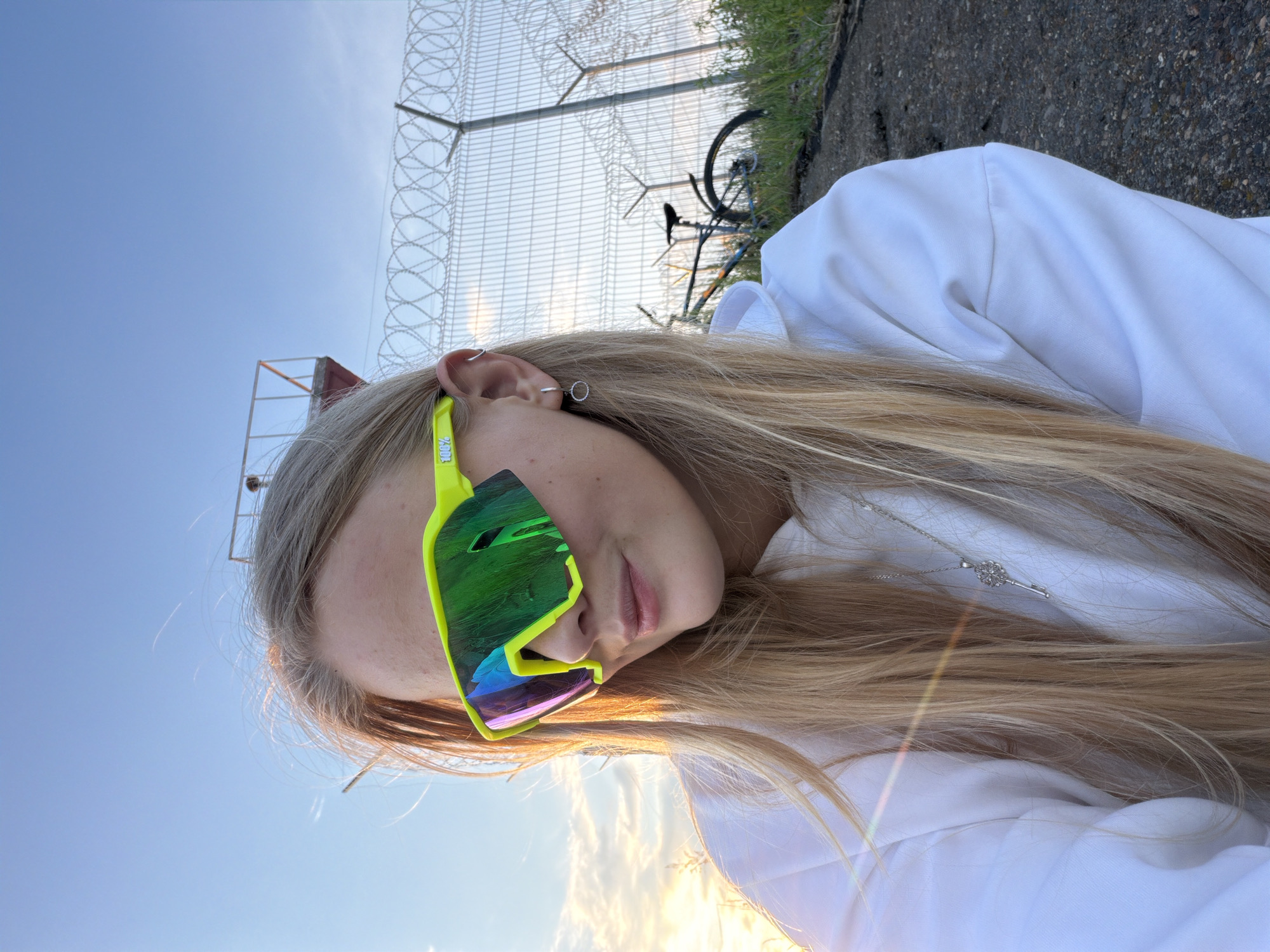 A young Caucasian female with long blonde hair is captured in a close-up, low-angle, outdoor shot. She is wearing a white hoodie, neon yellow sunglasses with mirrored lenses, and a silver necklace with a key pendant. A small hoop earring is visible in her left ear. The background reveals a clear blue sky with some wispy clouds, a chain-link fence with barbed wire, a distant structure resembling a watchtower, and a bicycle. The lighting suggests it is either sunrise or sunset, with a warm glow casting a lens flare across the image. The environment appears to be an outdoor area near a perimeter fence, possibly in an industrial or restricted zone within the city of Chaikovsky, Russia.