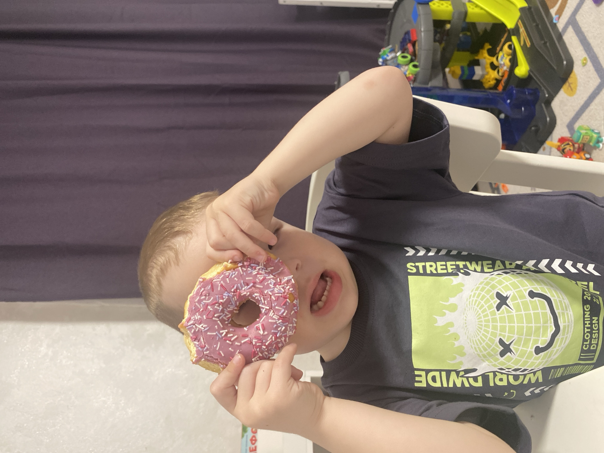 A child is holding a pink donut with sprinkles up to their eye. The child is sitting in a high chair. Behind the child is a purple curtain, a toy, and a patterned floor. The child is wearing a black t-shirt with a neon green graphic. There is nothing in the image to indicate that the location is Sochi, Russia.