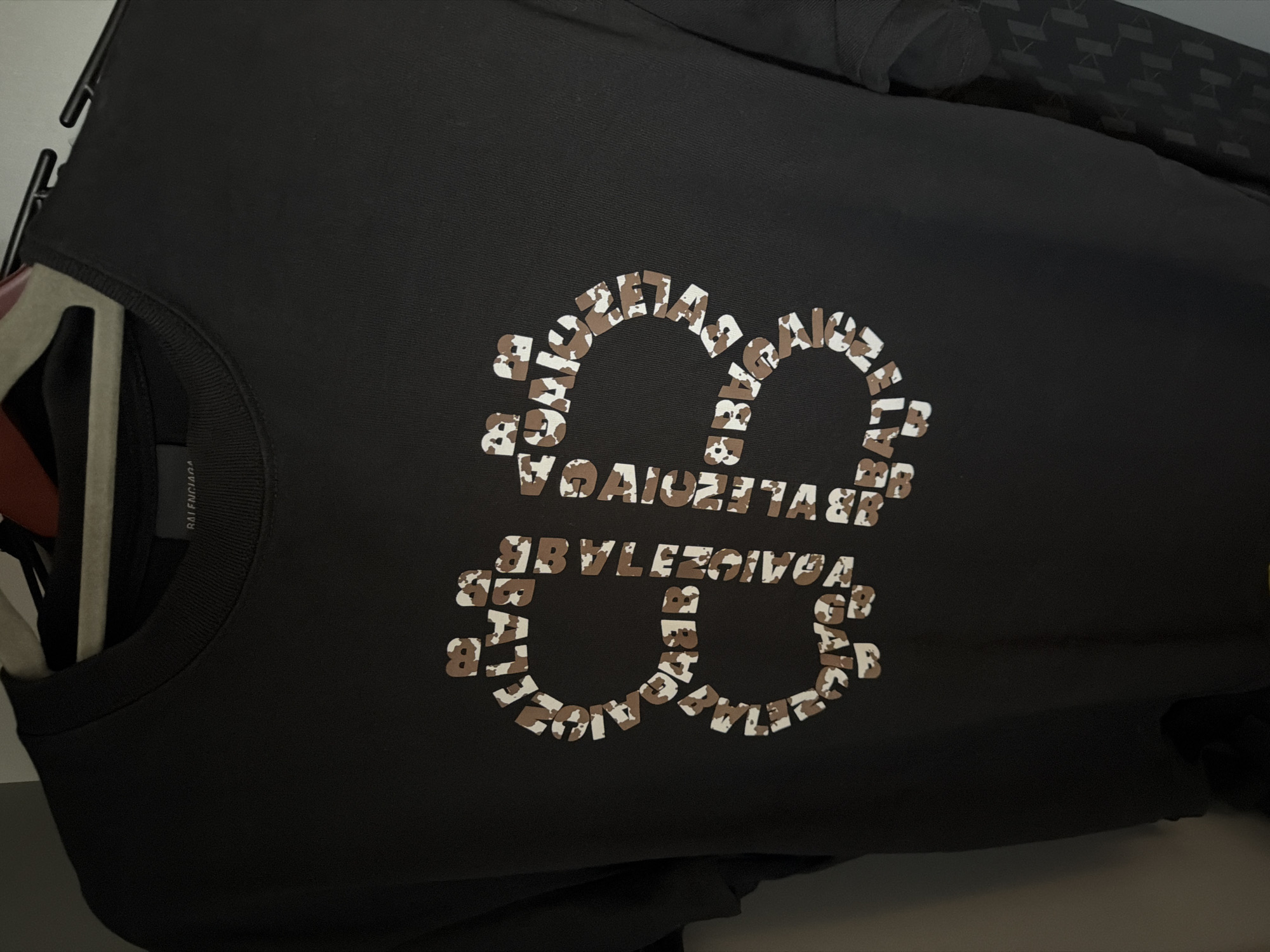 A black crewneck sweatshirt is depicted hanging on a light-colored wooden or plastic hanger with a dark metal hook, in the city of Sofia, Bulgaria. The sweatshirt features a ribbed collar, where a black label with "BALENCIAGA" printed in white capital letters is visible on the interior. The main graphic on the front of the sweatshirt is a large, stylized double-B logo, resembling a combined Bitcoin or dollar sign, rendered in a brown and off-white camouflage pattern. This pattern is formed by numerous smaller, repeating capital letters spelling "BALENCIAGA." The fabric of the garment appears to be a smooth, dark knit. To the lower right of the frame, a segment of another dark garment or fabric, embossed with a repeating "KL" monogram pattern, is partially visible. The background is a uniformly dark, indistinct surface, suggesting an indoor setting such as a closet or wall.