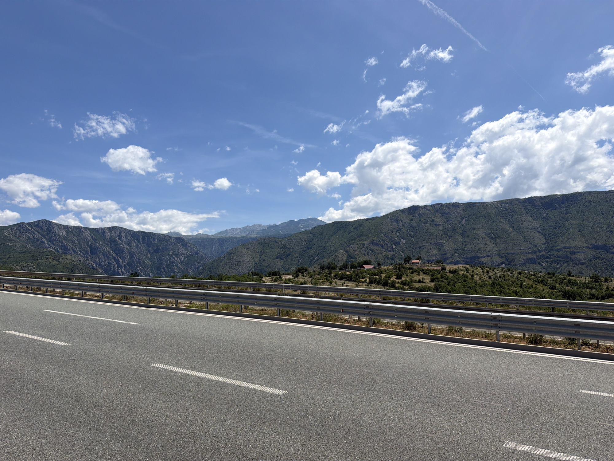 The image shows a highway with a metal guardrail in the foreground, bordered by a mountainous landscape. The sky is blue with scattered white clouds. Two red-roofed buildings are visible on a hill in the distance. The scene is likely located in Klopot, Montenegro, a region known for its scenic highways and mountainous terrain. There are no people or vehicles visible in the image.