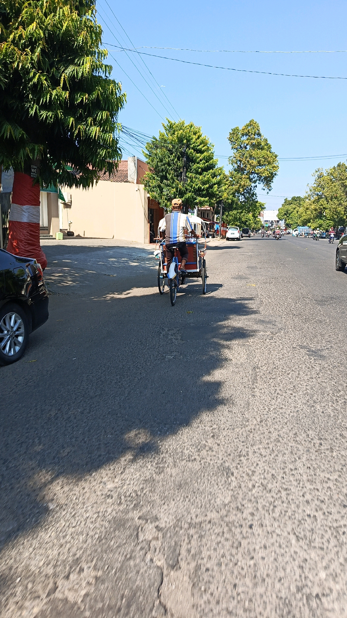 The image depicts a street scene in Nologaten, Indonesia, under a clear blue sky. The foreground is dominated by an asphalt road with visible textures and some minor cracking, casting a large shadow from unseen trees. To the left, a portion of a black car is visible. In the middle ground, a tricycle taxi, or "becak," is being ridden by a man wearing a striped shirt and a cap. The becak has a passenger compartment with red and blue accents and a white canopy. Behind the becak, other vehicles, including motorcycles and cars, are traveling on the road. Trees line the sides of the street, with one prominent tree on the left decorated with red and white striped fabric. Buildings with light-colored walls are visible on the left side of the road. The overall impression is that of a sunny day in a populated urban or suburban area.