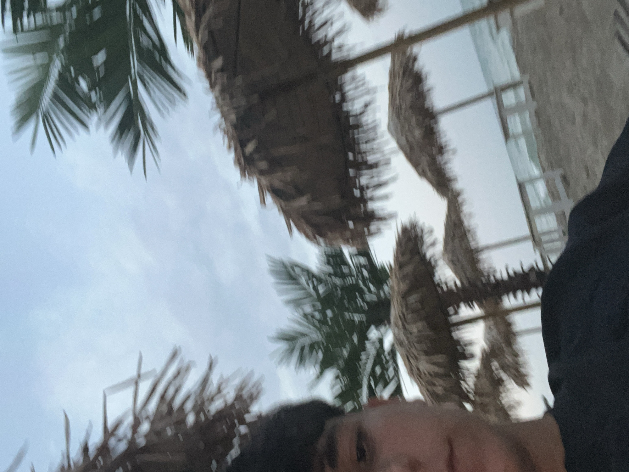 The image shows a close-up of a person's face on the left side, looking towards the camera. Behind the person, a beach scene unfolds. Several thatched umbrellas are visible, providing shade over what appear to be lounge chairs or tables. Palm trees with green fronds are scattered throughout the background, some in focus and others blurred. The ocean is visible in the distance with small waves lapping on the shore. The sky is overcast and appears to be either early morning or late afternoon. The sand is light brown. The location is the Black Sea coast in Obzor, Bulgaria, indicated by the specific architecture of the umbrellas and the general setting of a beach resort town. The image is somewhat blurry, suggesting it was taken on a moving subject or in low light conditions.