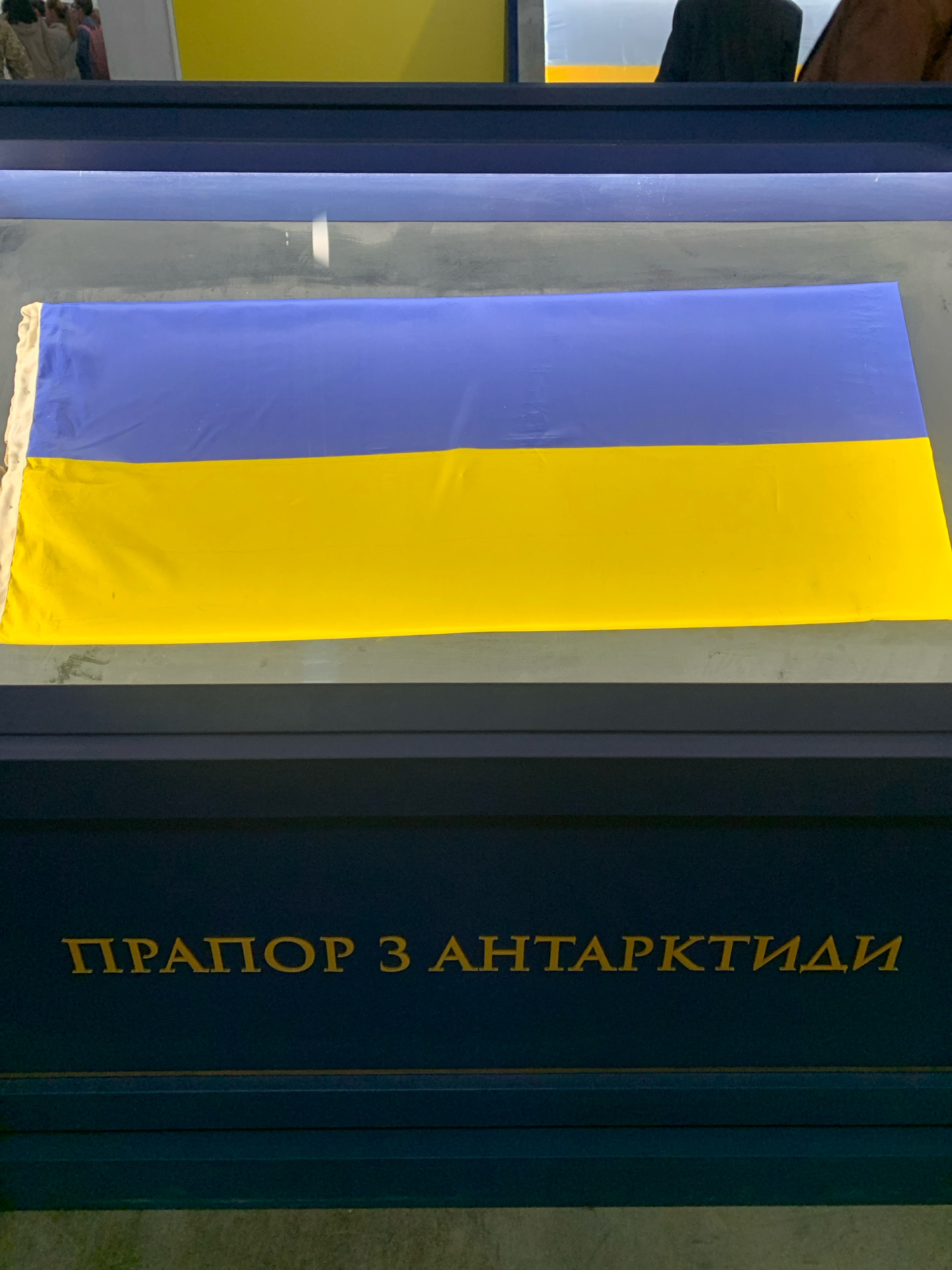 The image shows a Ukrainian flag displayed in a dark blue exhibit case with gold lettering. The flag, which is divided horizontally into blue and yellow sections, is laid out flat and appears to be behind a glass or transparent barrier. The gold text on the case reads "ПРАПОР З АНТАРКТИДИ," which translates to "FLAG FROM ANTARCTICA." The background of the exhibit case is dark, with a horizontal gold line above the text. The lighting in the exhibit suggests an indoor setting, possibly a museum or a public exhibition space. The overall impression is one of a historical or symbolic display. The specific location in Kyiv, Ukraine, cannot be determined from the image alone.