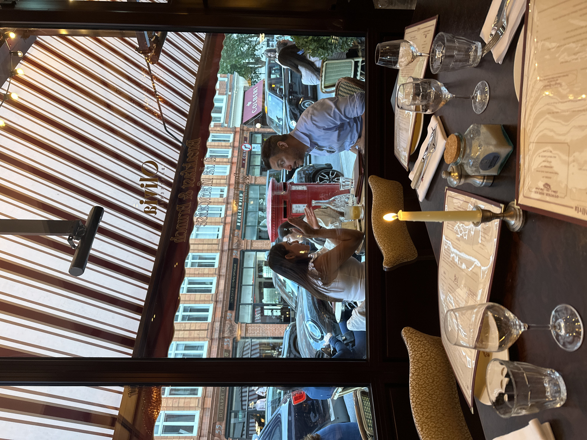 A man and a woman are seated at a table inside a restaurant, visible through a large window that overlooks a street scene. The man, wearing a light blue collared shirt, is looking to his left, with his hands resting on the table. The woman, wearing a white top, is looking and gesturing towards the man, seemingly engaged in conversation. On the table in front of them are glasses, a bottle of water, and menus. Outside the window, a red post box and parked cars are visible, along with buildings and a striped awning over the restaurant. The foreground of the image shows a table with place settings, including menus, cutlery, glassware, and a lit candle in a candlestick holder. The overall lighting suggests it is evening or dusk.