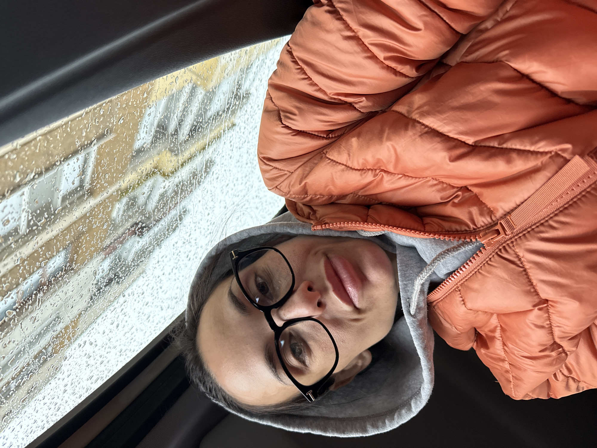 A woman is depicted from an upside-down perspective. She is wearing black-framed glasses and a gray hooded sweatshirt, partially covered by an orange puffer jacket. The scene suggests she is inside a vehicle, as evidenced by a rain-streaked window and a car's interior frame. The environment visible through the window appears to be an urban setting, possibly a street with buildings. There is no indication to confirm the location is Pleven, Bulgaria, based on the visual elements alone.