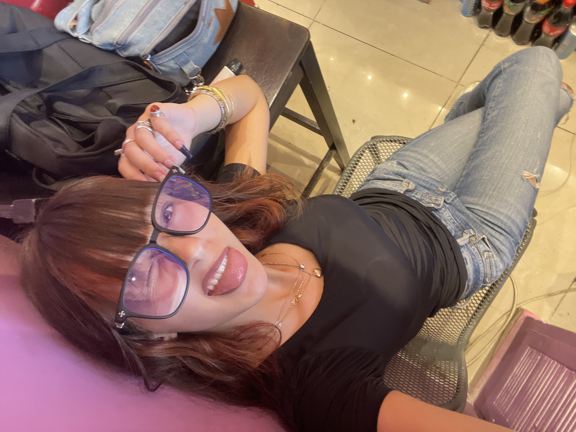 A young woman is captured from a high-angle, selfie-style perspective, sitting on a metal chair in what appears to be an indoor public space with tiled flooring. She is wearing a black t-shirt, blue jeans with a distressed knee, and large, square-framed glasses. Her tongue is sticking out, and she is winking. She has long, dark hair with reddish highlights and is adorned with multiple rings and bracelets. Behind her, a dark backpack and a denim bag are visible, suggesting she might be traveling or at a transit point. The background consists of a pink wall on the left and a wooden table on the right, holding some items. The lighting suggests an indoor environment, possibly a cafe or lounge area. The location is identified as Raanana, Israel.