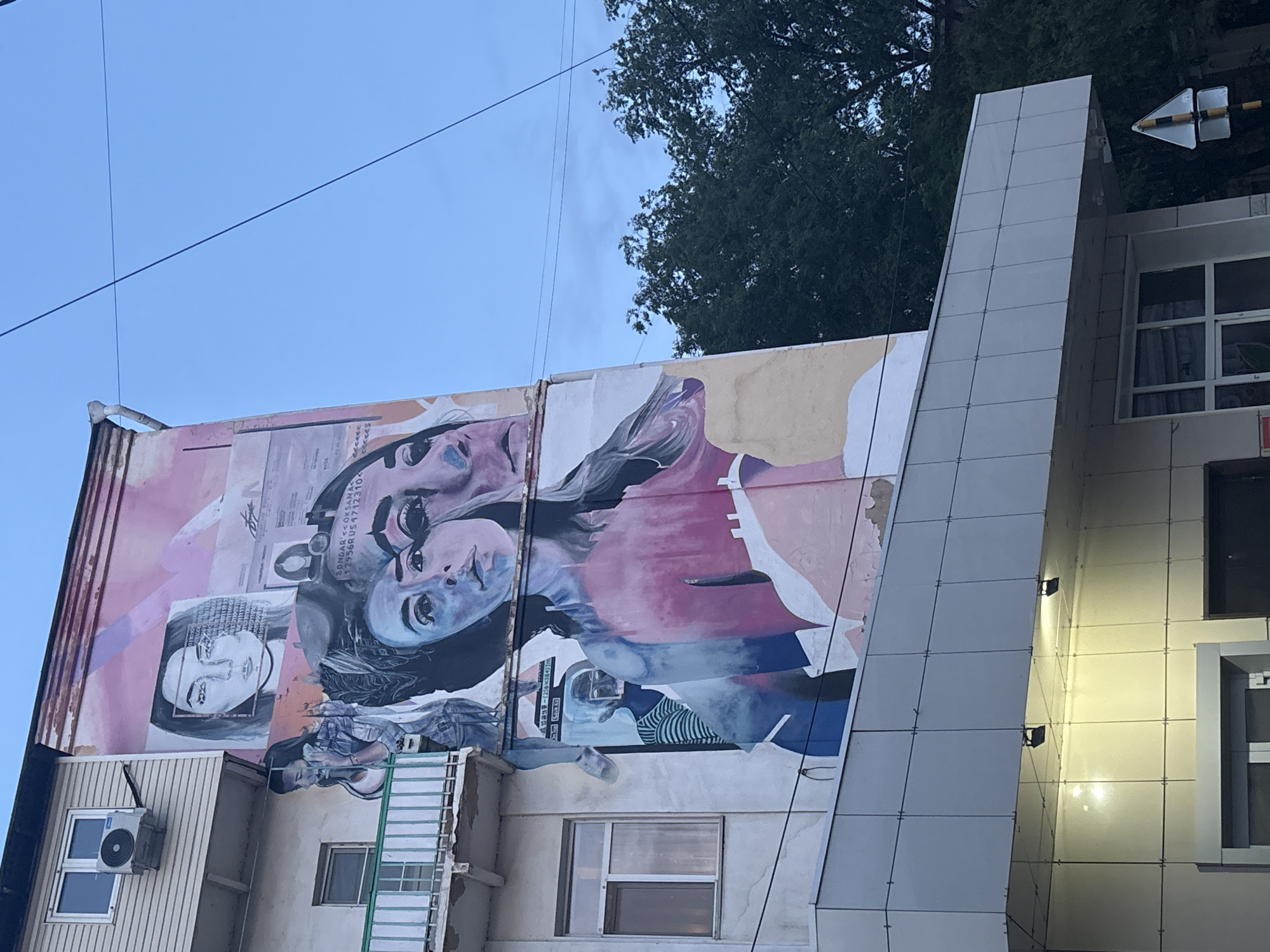 The image shows a mural on the side of a multi-story building in Astrakhan, Russia. The mural is a collage of portraits of women and appears to be in an abstract and stylized manner. The building itself is light-colored with multiple windows and a balcony on one side. There is an air conditioning unit mounted on the exterior wall above the balcony. A modern facade with lights is visible at the bottom of the building. Trees are visible behind the building on the right side, and the sky is visible in the upper portion of the image. The overall scene depicts urban art on a building in a city setting.