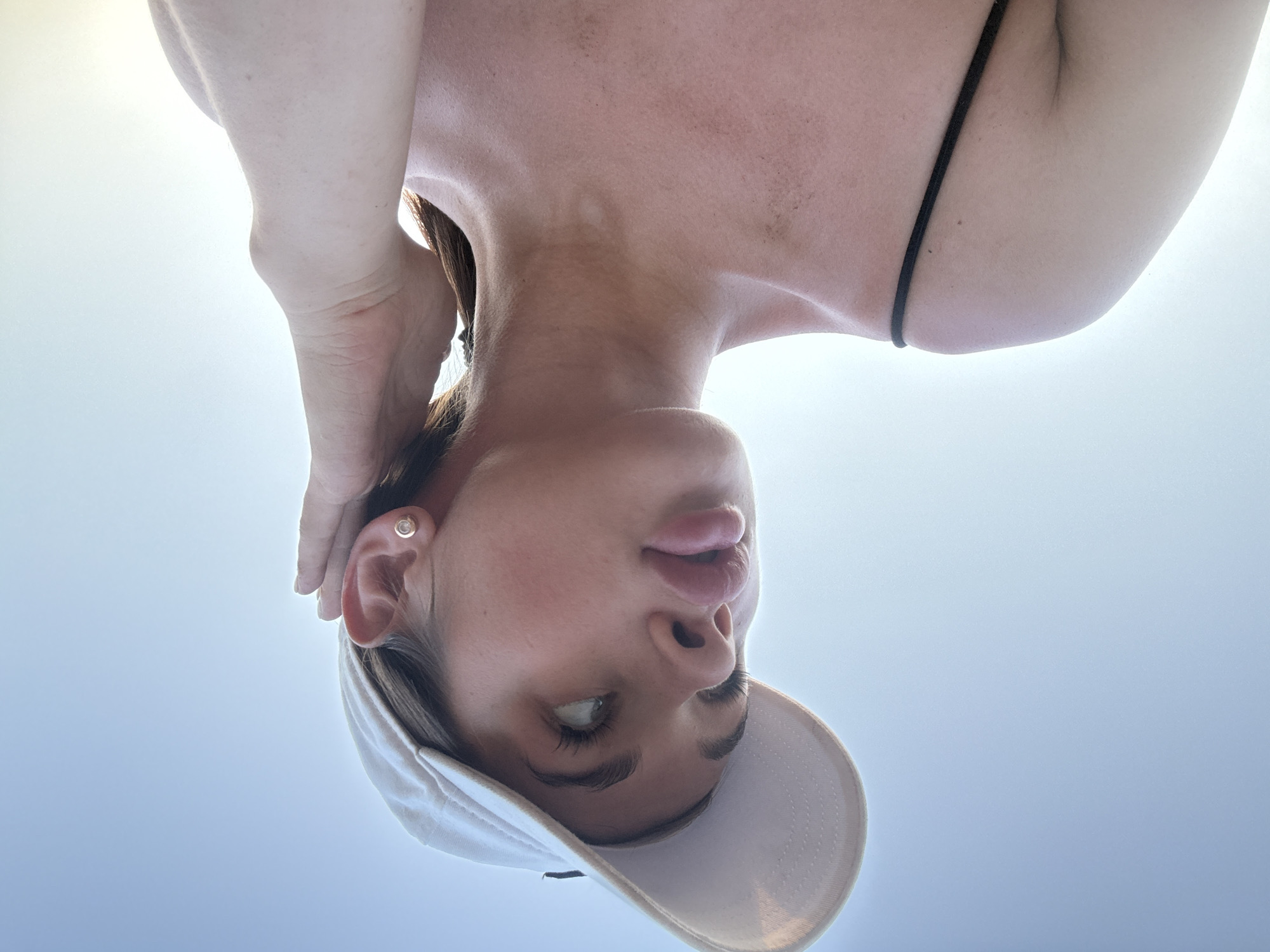A close-up, low-angle, waist-up shot shows a young woman with full lips, wearing a white baseball cap and a black spaghetti strap top. She has a small stud earring in her right ear and is touching her hair with her right hand. Her head is tilted upwards and to the left, looking away from the camera. The background is a clear, pale blue sky, suggesting an outdoor setting, possibly during daylight hours. The lighting indicates the sun is behind and to the left of the subject, creating a soft glow. There are no visible landmarks or features that can definitively place this image in Lobanovo, Russia.