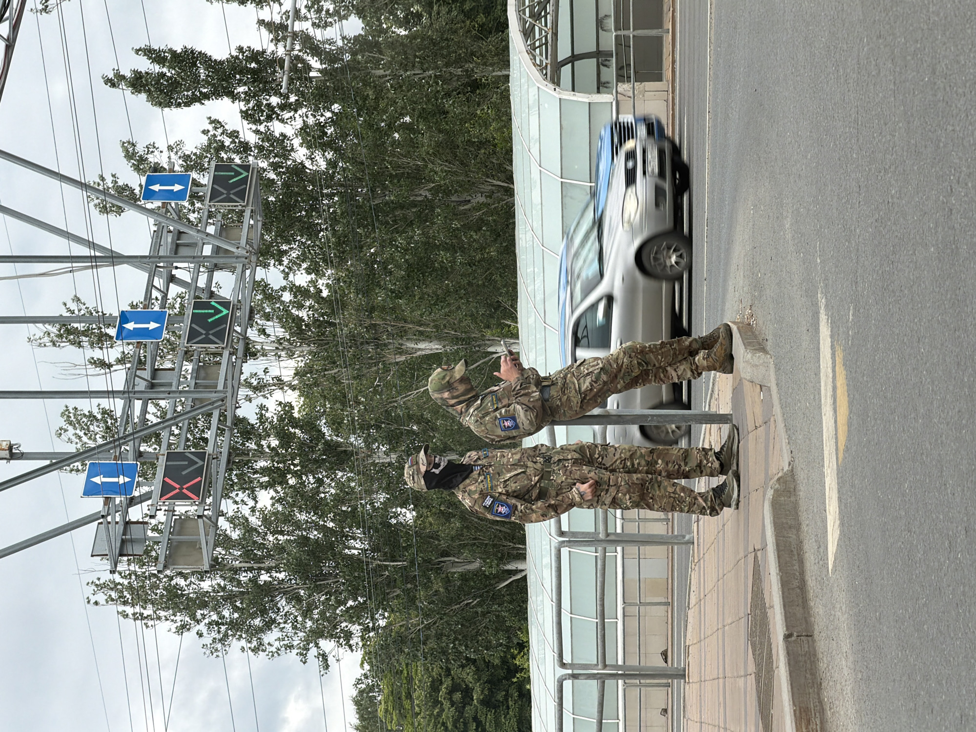 This image captures an outdoor street scene under an overcast sky, purportedly located in Samara, Russia. The foreground shows an asphalt road with a partial yellow lane marking, adjacent to a paved sidewalk separated by a concrete curb.
Two individuals are present on the sidewalk, both dressed in multi-camouflage military uniforms and tactical footwear. The individual on the left stands upright, wearing a camouflage cap, a black balaclava with a white skull design, and a uniform displaying patches. A blue shield-shaped patch with a white emblem and Cyrillic text (partially visible, including "ШТУРМАН" above it) is on their right sleeve, and a similar patch is on their left sleeve. A tattoo is visible on the back of their left hand. The individual on the right leans against a silver metal barrier, looking down at a smartphone held in both hands. They are also wearing a camouflage cap and a multi-cam uniform with a blue shield-shaped patch visible on their left sleeve.
Behind the metal barrier, an arched, translucent-roofed structure, possibly a pedestrian cover or bus stop, extends horizontally. Vehicles are visible on the road; a silver sedan is in motion from right to left, partially obscured by blur, followed by a blue vehicle.
Overhead, a metal gantry spans the road, supporting multiple traffic signals and blue directional signs. From left to right, the traffic signals display a red 'X', a green downward arrow, another green downward arrow, and a red 'X'. Above these signals, four blue square signs each feature a white double-headed vertical arrow, indicating reversible lane operation. Numerous electrical or utility wires crisscross the upper portion of the frame. The background is dominated by tall, slender trees with green foliage, suggesting a wooded or park-like area.