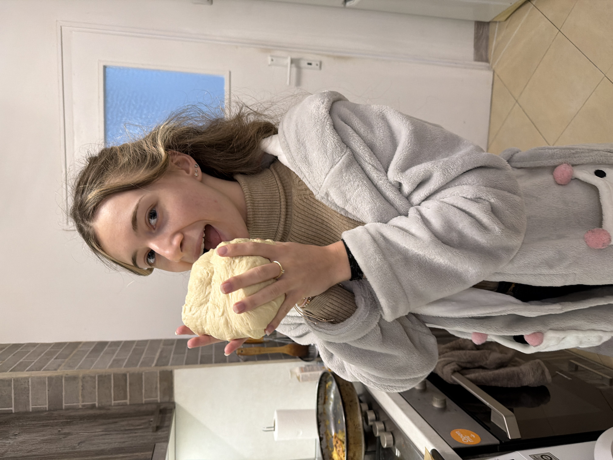 A young woman in a light gray, plush robe with pink pom-poms is holding a ball of dough up to her open mouth, seemingly about to take a bite. She is also wearing a tan ribbed shirt or sweater. The scene appears to be inside a kitchen. A countertop runs along the bottom left, with a light gray tile backsplash. In the background is a white door with a rectangular glass panel. An oven with visible burner knobs is visible on the bottom right. A towel roll is on the counter behind the woman. The woman is fair-skinned with light brown hair. The image is rotated counterclockwise. Location is undetermined and the city of Plovdiv, Bulgaria is not confirmed.