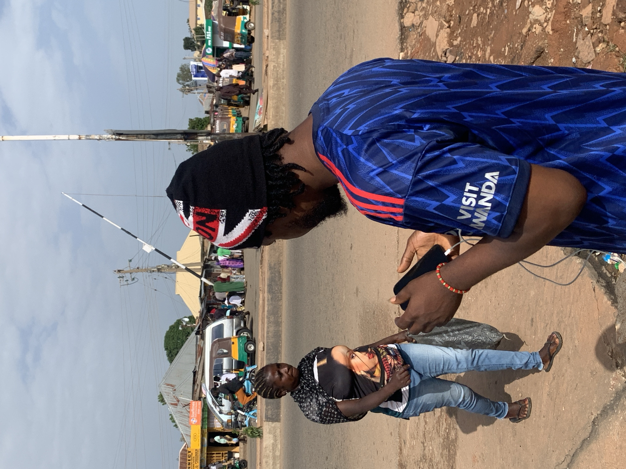 In Gura Top, Nigeria, an overcast sky looms above a bustling street scene. In the foreground, a man wearing a blue jersey with red accents and a black beanie adorned with the Union Jack is engrossed in his mobile phone. To his left, a woman walks with a black plastic bag in her right hand, sporting a black and white patterned scarf over a t-shirt featuring a portrait of a woman. She wears blue jeans and dark flip-flops. The background reveals a street lined with shops and vehicles, including a white van and several auto-rickshaws. Pedestrians are visible in the distance, navigating the marketplace. A railway crossing barrier stands prominently in the mid-ground. The ground is a mix of dirt and pavement, with scattered rocks and debris.