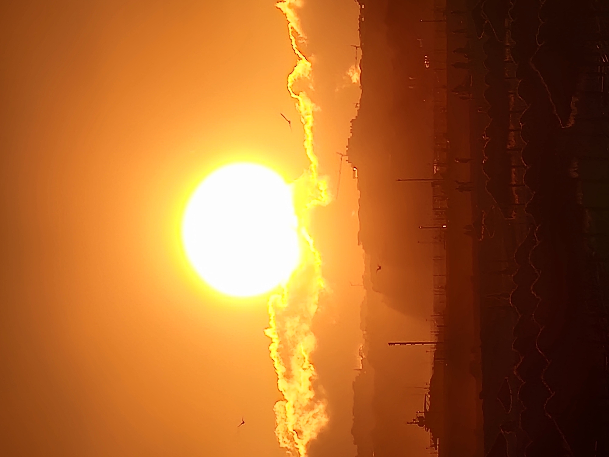 The image depicts a sunset over Quarteira, Portugal, with the sun prominently featured in the upper center of the frame. The sun is a bright, glowing orb, casting a warm, orange hue across the sky and illuminating the clouds below. These clouds are thick and textured, with their edges highlighted by the intense sunlight, creating a dramatic contrast.
In the mid-ground, a silhouetted cityscape stretches across the horizon. Buildings and structures are dimly visible, suggesting an urban or developed area. The outlines of cranes can be discerned among the buildings, hinting at ongoing construction or industrial activity. A vessel, possibly a ship or ferry, is also visible on the left side of the mid-ground, partially obscured by the haze.
The foreground is dominated by rows of thatched beach umbrellas, forming a dense pattern that stretches across the bottom of the image. These umbrellas are dark silhouettes against the brighter sky, suggesting they are either closed or unoccupied. Scattered figures of people are faintly visible in the mid-ground, walking or standing, adding a sense of scale and human presence to the scene. A few birds are silhouetted against the sky, adding a dynamic element to the composition. The overall atmosphere is one of a serene evening at a coastal location.