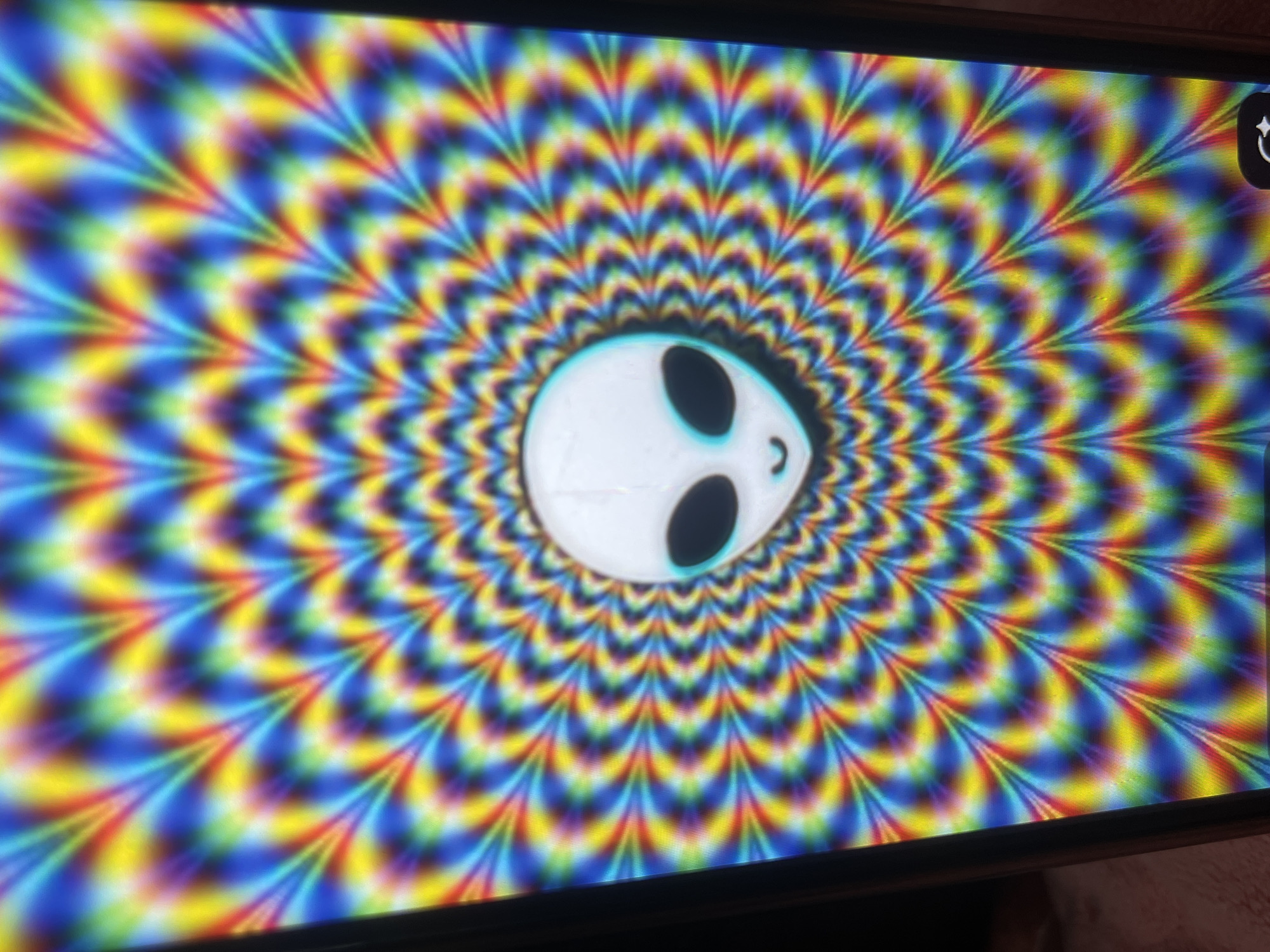 The image displays a close-up, vertically oriented shot of a smartphone screen. The screen exhibits a psychedelic pattern of concentric, radiating waves of color, primarily featuring blues, yellows, reds, and purples. Centered within this pattern is a stylized, cartoonish alien face with large, black oval eyes and a small, smiling mouth. The alien's head is a pale white, outlined with a faint blue glow. The image does not provide any context or details related to Bishkek City, Kyrgyzstan, as it is solely focused on the digital display of the phone. There are no visible people, objects, or environmental elements outside of the phone's screen.