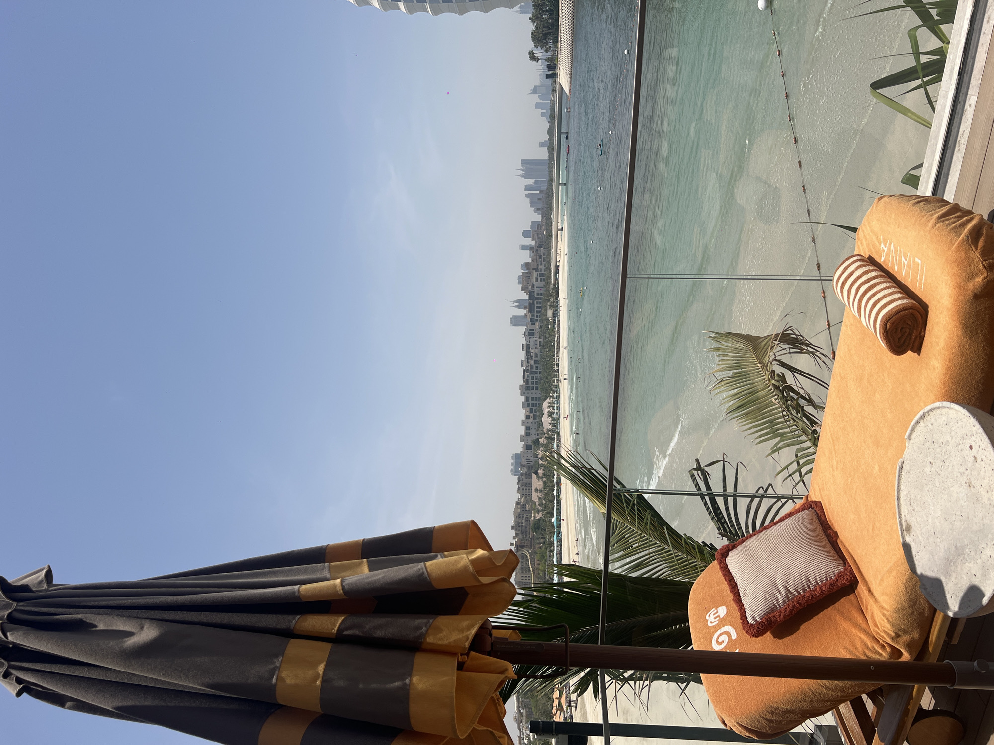 The image depicts an elevated view overlooking a beach in Dubai, United Arab Emirates, likely from a hotel balcony or rooftop. The view is focused on the beach and coastline, which features light-colored sand and turquoise water. Buildings, including residential and commercial structures, line the beach. The sky is clear and blue.
In the foreground is an outdoor lounge area. A sun umbrella, primarily grey with gold accents, is positioned to provide shade. A sun lounger with a cushion, a striped towel, and a small table are visible. A palm tree's leaves are visible between the lounger and the beach. A clear glass barrier separates the lounge area from the open view of the beach.