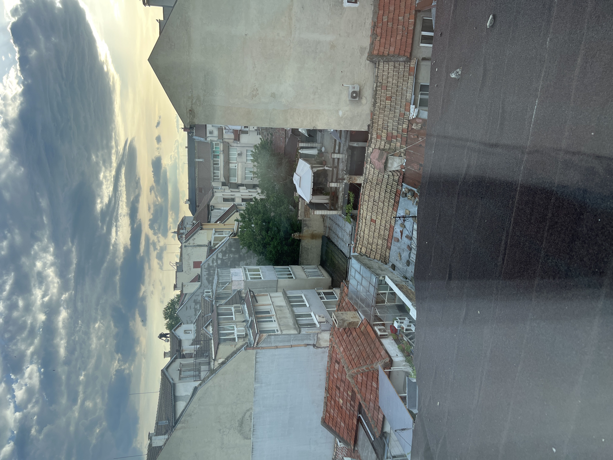 The image depicts a rooftop view in Varna, Bulgaria, featuring a mix of residential buildings with varied architectural styles and ages. The buildings are tightly packed, with visible red-tiled roofs and white or beige facades. Some buildings have balconies and windows with visible air conditioning units. A green tree is positioned in the courtyard between the buildings. The sky is visible in the upper left corner with a cloudy overcast condition. In the lower right corner is a flat, dark rooftop, possibly from the building where the photo was taken.