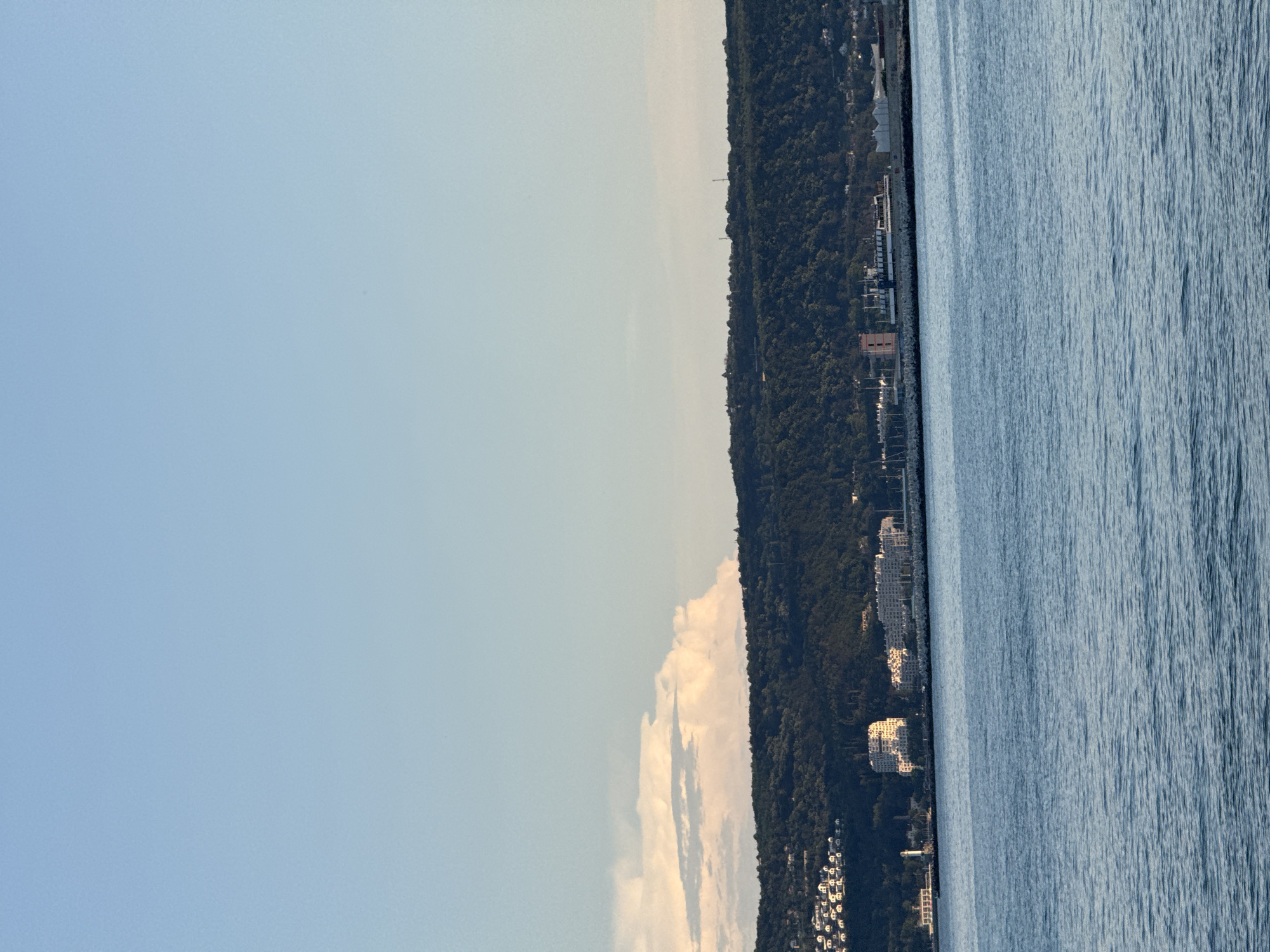 An eye-level shot shows a wide expanse of calm, blue water in the foreground, leading to a distant, verdant hillside dotted with buildings and a harbor. The sky above is a clear, pale blue, with a few scattered, fluffy white clouds to the left. The hillside is covered in dense, dark green trees, and a cluster of white buildings with red-tiled roofs is visible on its lower slopes. Near the water's edge, several larger, modern-looking white buildings stand, some with distinctive geometric designs. A lighthouse and a red-and-white tower are also present along the waterfront, suggesting a port or industrial area. The overall impression is of a coastal city scene under a bright sky.