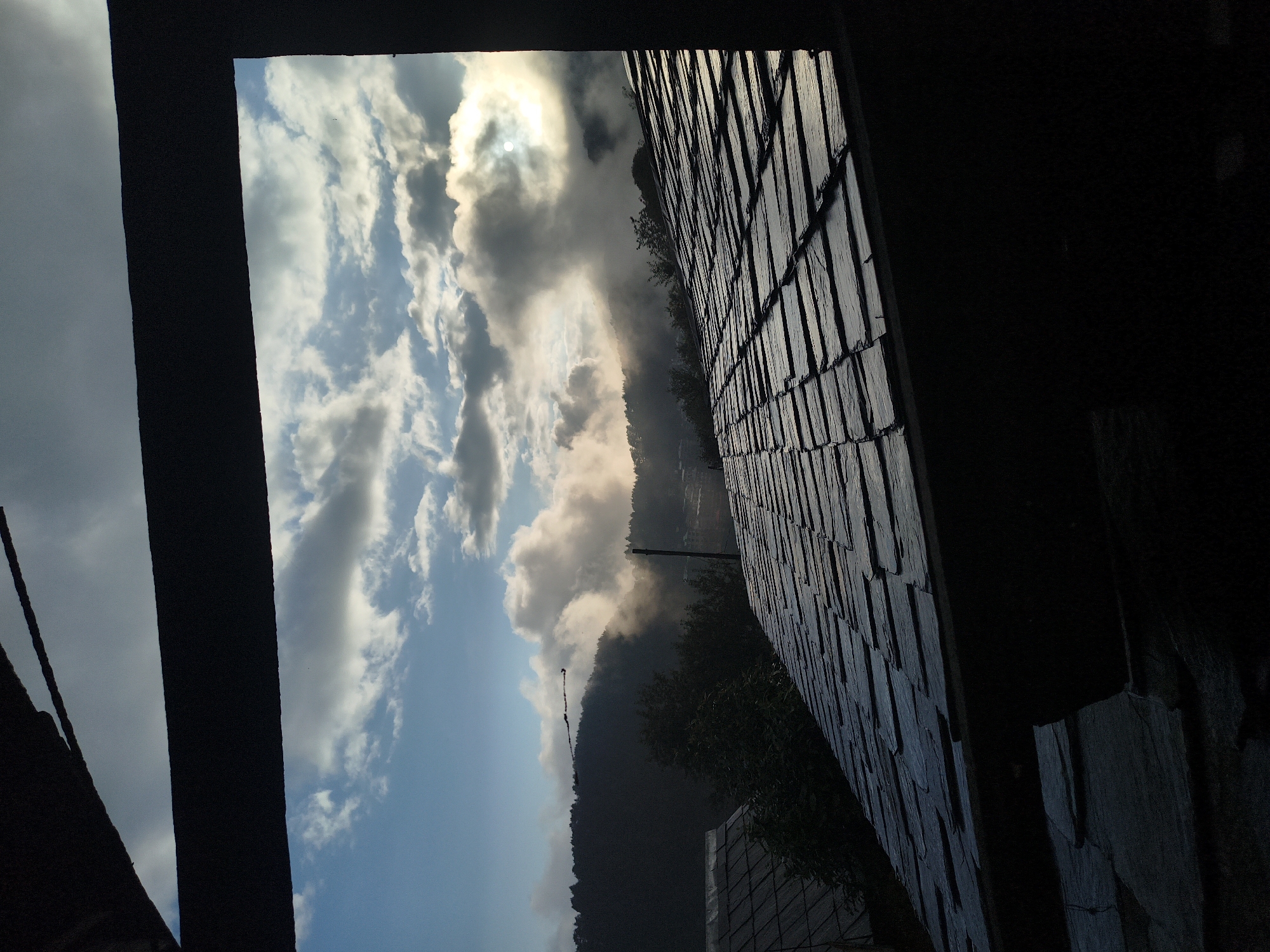 The image is a photograph taken from an interior, looking out through a dark, silhouetted frame onto a landscape under a cloudy sky. The foreground is dominated by the dark shapes of what appear to be roof structures covered in slate tiles. The roof tiles are dark gray and appear weathered. Beyond the roofs, a treeline of dark green trees forms a silhouette against a bright, partly cloudy sky. The clouds are white and puffy in some areas, with darker, more dramatic formations in others. The sun is partially obscured by clouds, casting a diffused, bright light on the scene. In the middle ground, a distant hillside is visible, also covered in trees and possibly some structures. There are no people visible in the image. The overall impression is of a mountainous or hilly region, possibly at dawn or dusk due to the quality of light. The location is specified as Haini, India.