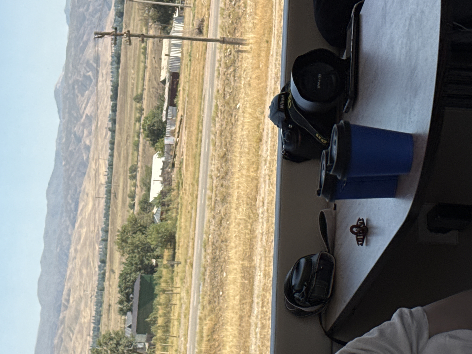 A camera and two blue cups are on a table in front of a view of mountains. The camera is a Nikon. The cups are paper and have black lids. The table is brown and appears to be made of wood. In the background, there are mountains and some buildings. The sky is blue. The location is Sasyk-Bulak, Kyrgyzstan.