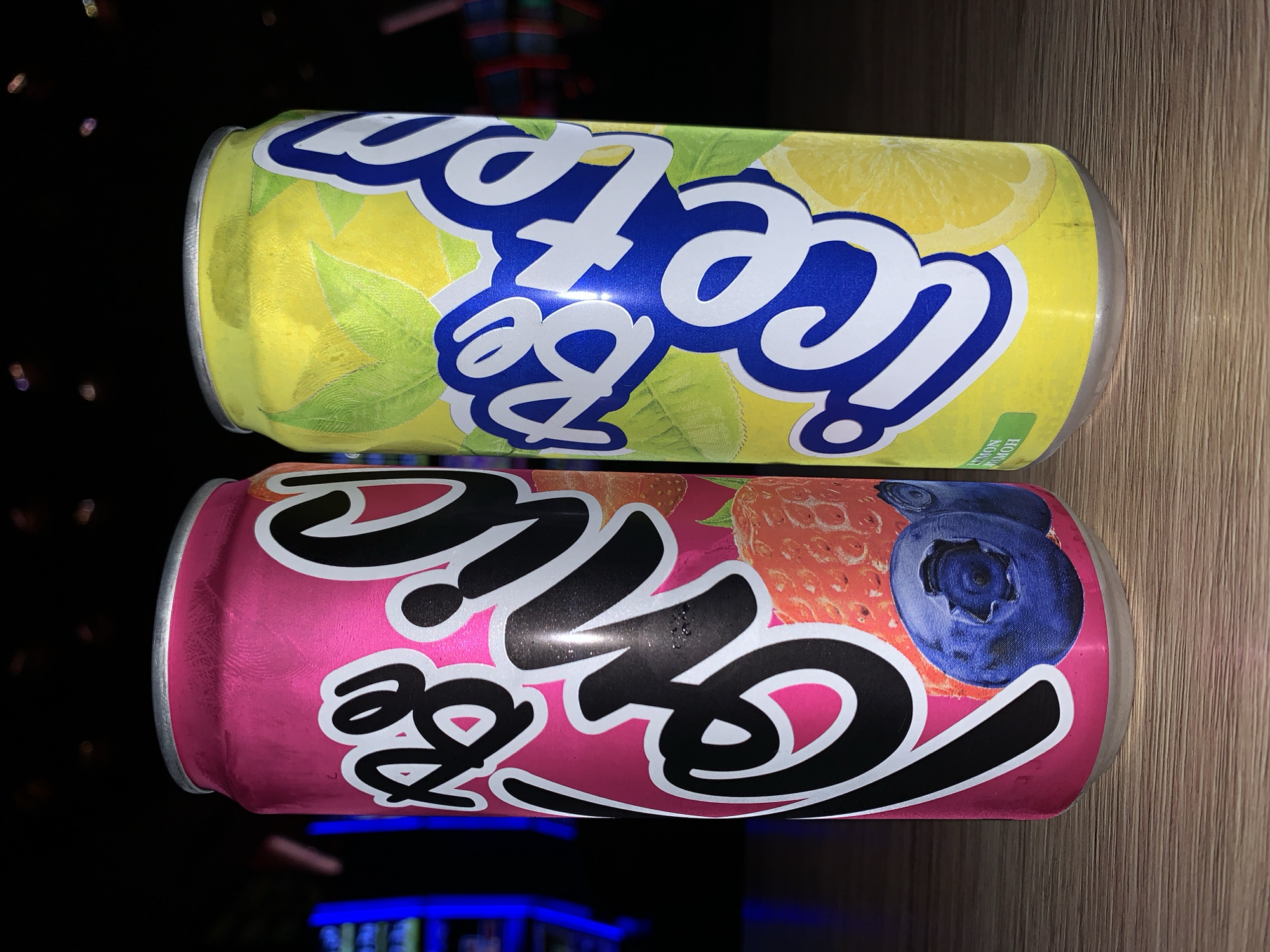 Two cans of "Be Ice Tea" are placed side-by-side on a wooden surface. The can on the left is pink and features strawberries and blueberries on its label. The can on the right is yellow and displays lemons and green leaves. The background is dark, with blurred colorful lights suggesting an indoor environment, possibly a bar or arcade. The cans are condensation-covered, indicating they are cold. No people are visible in the image. The scene is not identifiable as being in Burgas, Bulgaria.