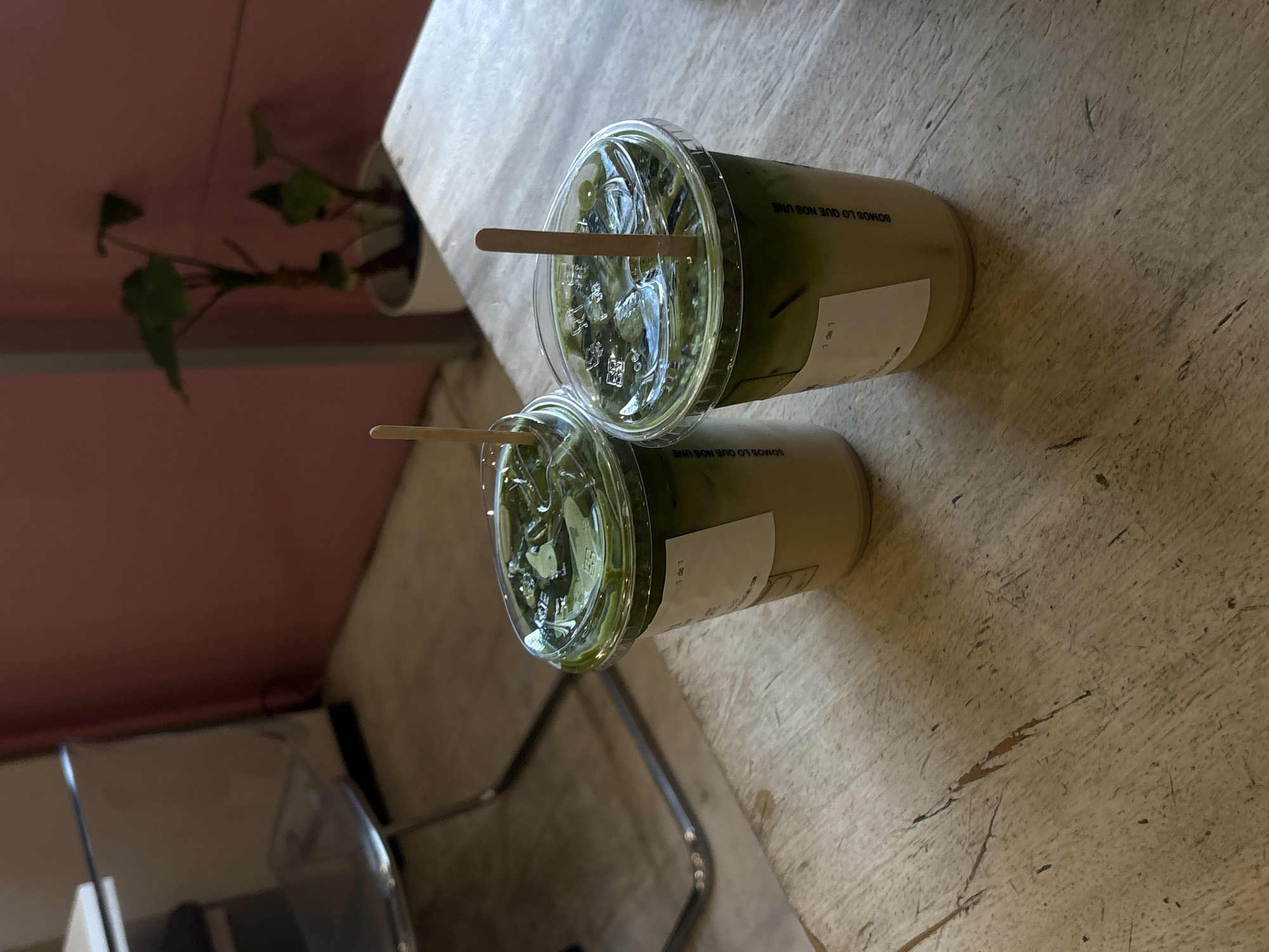 The image shows two clear plastic cups filled with a light green liquid, likely matcha lattes, on a light-colored, possibly wooden, surface with a worn texture. Each cup has a lid and a wooden stir stick. The cups bear labels with the text "1 de 1 SOMOS LO QUE NOS UNE". In the background, there is a glimpse of a plant in a white pot and a pink wall. The overall setting appears to be a cafe or similar establishment, potentially in Puebla, Mexico, based on the Spanish text on the cups. A chair can be seen in the lower left corner.