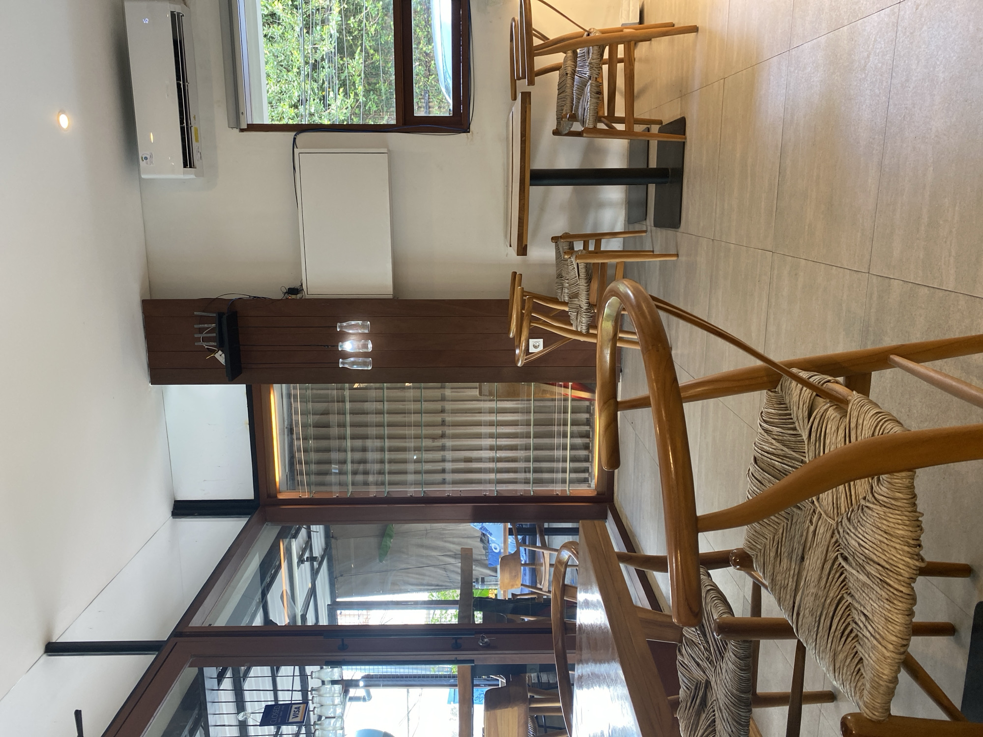 The image captures an interior view, likely a restaurant or cafe, in North Kuta, Indonesia. The scene features a dining area with wooden chairs having woven seats, arranged around tables with dark bases. Natural light streams through a window, showcasing a view of greenery outside. A glass sliding door provides a glimpse into an outdoor space. The interior wall is adorned with a minimalist design element - a dark wooden panel with glass bottles. An air conditioning unit is mounted on the wall above. The floor is tiled in neutral shades. The overall atmosphere appears relaxed and contemporary.