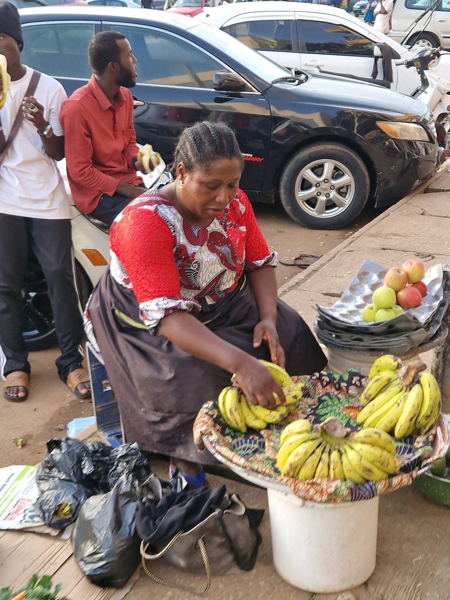 A full shot shows a street scene with people and produce in Jos, Nigeria. In the foreground, a woman with dark skin and braided hair is seated behind a display of bananas, dressed in a red and white patterned top and a dark skirt. Her hands are arranging a bunch of bananas on a colorful cloth-covered surface. To her left, a stack of items, including what appears to be a metal tray with apples, is visible. To the left of the woman, a man wearing a red shirt and black pants sits, holding a banana. Next to him, another man in a white t-shirt and black pants also holds a banana. In the background, a dark sedan is parked, with its wheels and part of the car visible. The ground is a mix of dirt and concrete, with various bags and items scattered around the scene, suggesting a market or roadside stall environment.
