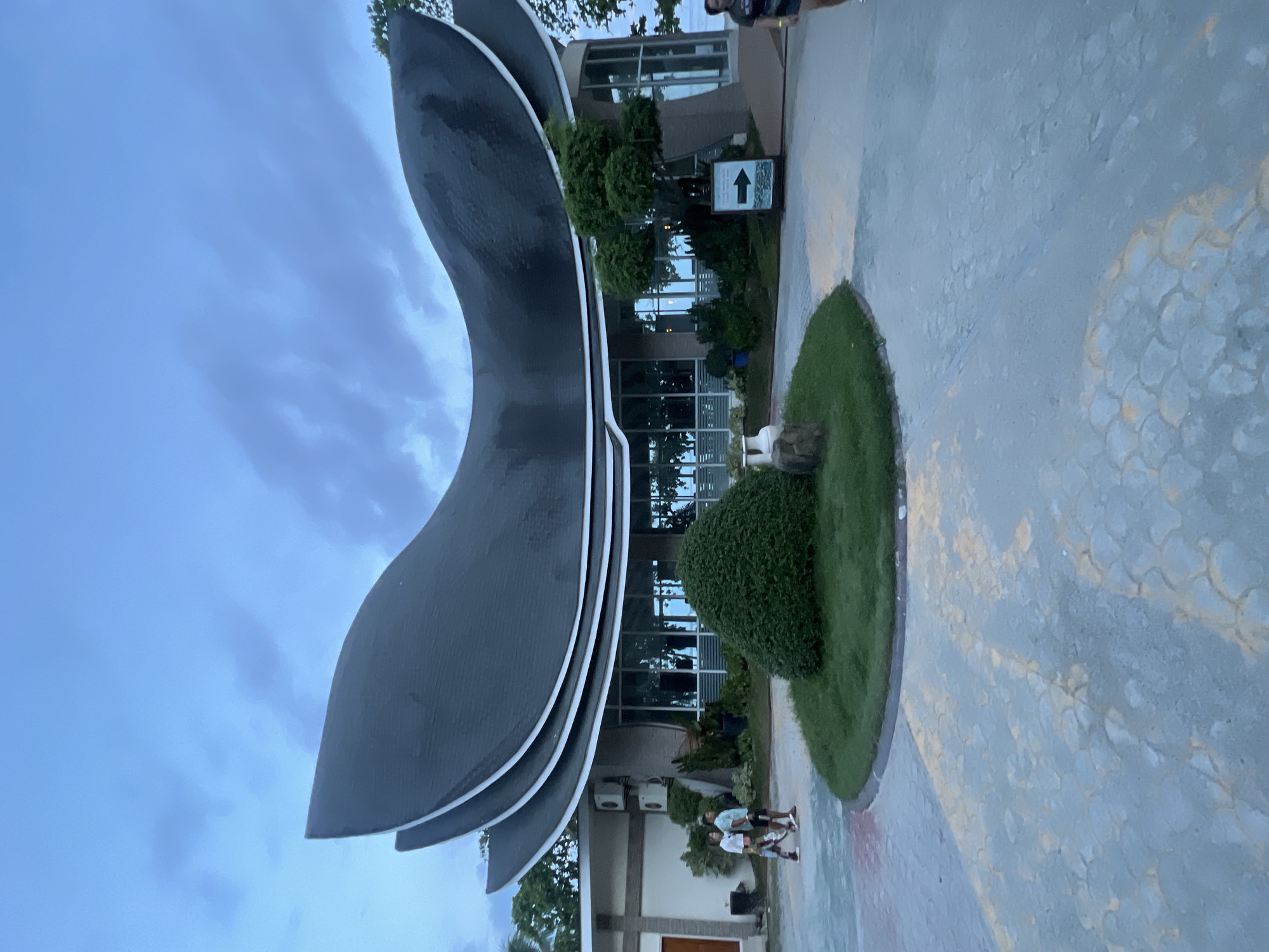 The image captures the Municipality of Banaybanay Hall in Davao Oriental, Philippines. The hall features a modern architectural design with curved, gray-toned structures lined with white trim. Large glass windows are visible, reflecting the outdoor environment.
On the right side of the image, a circular patch of green grass and a stone pathway is visible. A signpost with an arrow is present.
Two individuals, appearing to be children, are seen walking on the patterned pavement in front of the building. The sky above is overcast with a mix of blue and gray clouds.