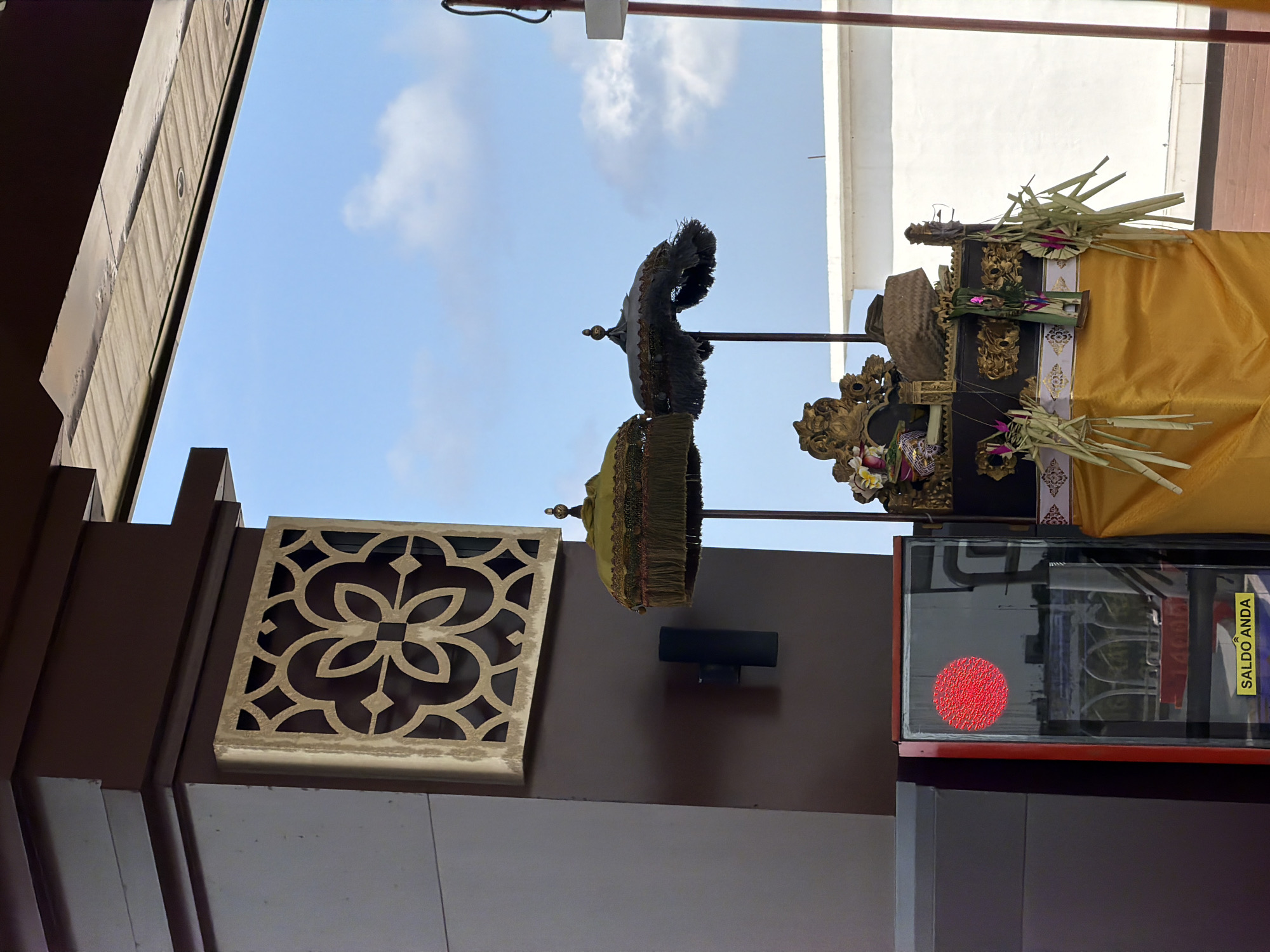This image depicts an outdoor scene, likely a temple or cultural site in South Denpasar, Indonesia. The sky is visible, suggesting daytime with some clouds. The foreground is dominated by dark brown architectural elements and a decorative lattice panel with a floral motif.
In the mid-ground, traditional Balinese offerings are prominently displayed. These include ornate umbrellas, one dark and fringed, and another in a muted green with decorative tassels, both appearing to be part of a procession or ceremony. They are placed on a raised platform draped with a vibrant yellow cloth, adorned with gold trim and what appear to be woven palm leaf decorations. The overall arrangement suggests a religious or ceremonial context.
Further to the right, a glass-fronted display case is visible. Inside, a red circular pattern is illuminated, possibly an indicator or part of an exhibit. A yellow sticker with text is affixed to the right side of the display case. The text on the sticker is "SALDO ANDA", which translates from Indonesian to "YOUR BALANCE". This hints at the possibility of a transactional or service element within the cultural setting, perhaps related to donations or ticket sales.
The architecture suggests a blend of modern and traditional elements, with the dark brown structures contrasting with the bright yellow fabric and the intricate lattice work. The lighting indicates it is likely daytime, with diffused sunlight illuminating the scene. The presence of the ornate umbrellas and offerings strongly points to a cultural event or ongoing religious practice.
