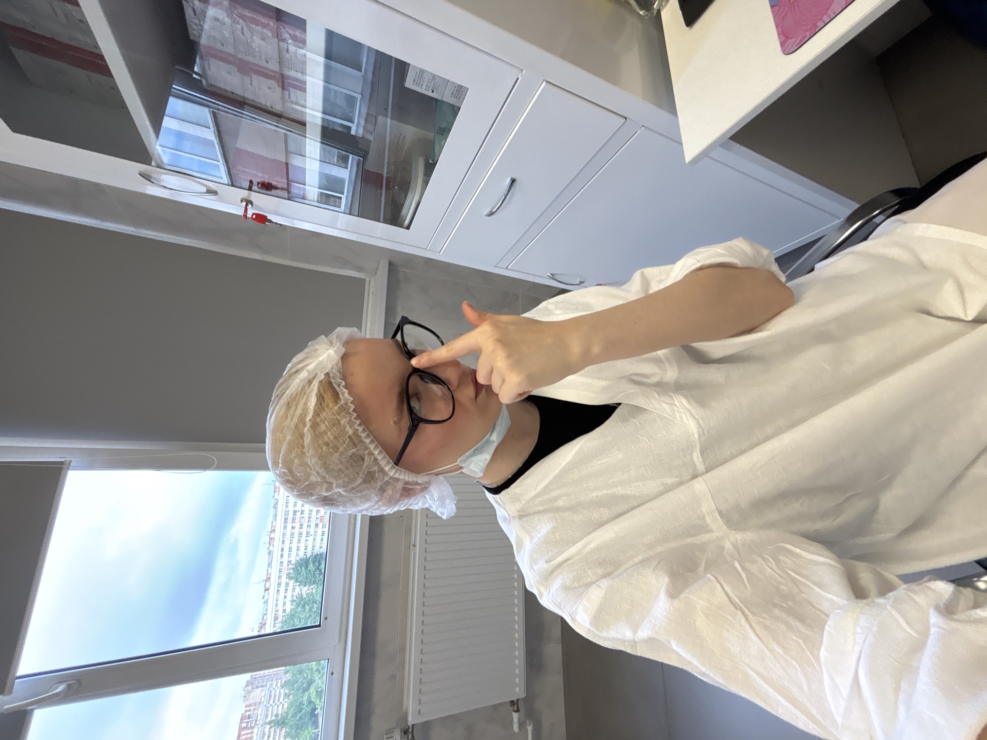 A person wearing a white disposable medical gown, a hairnet, and glasses is sitting in a room. They are touching their glasses with their right index finger and thumb. A light blue surgical mask is hanging around their neck. The room appears to be a medical or laboratory setting, with a white cabinet with glass doors filled with items visible in the background. A window shows a cloudy sky and buildings outside.